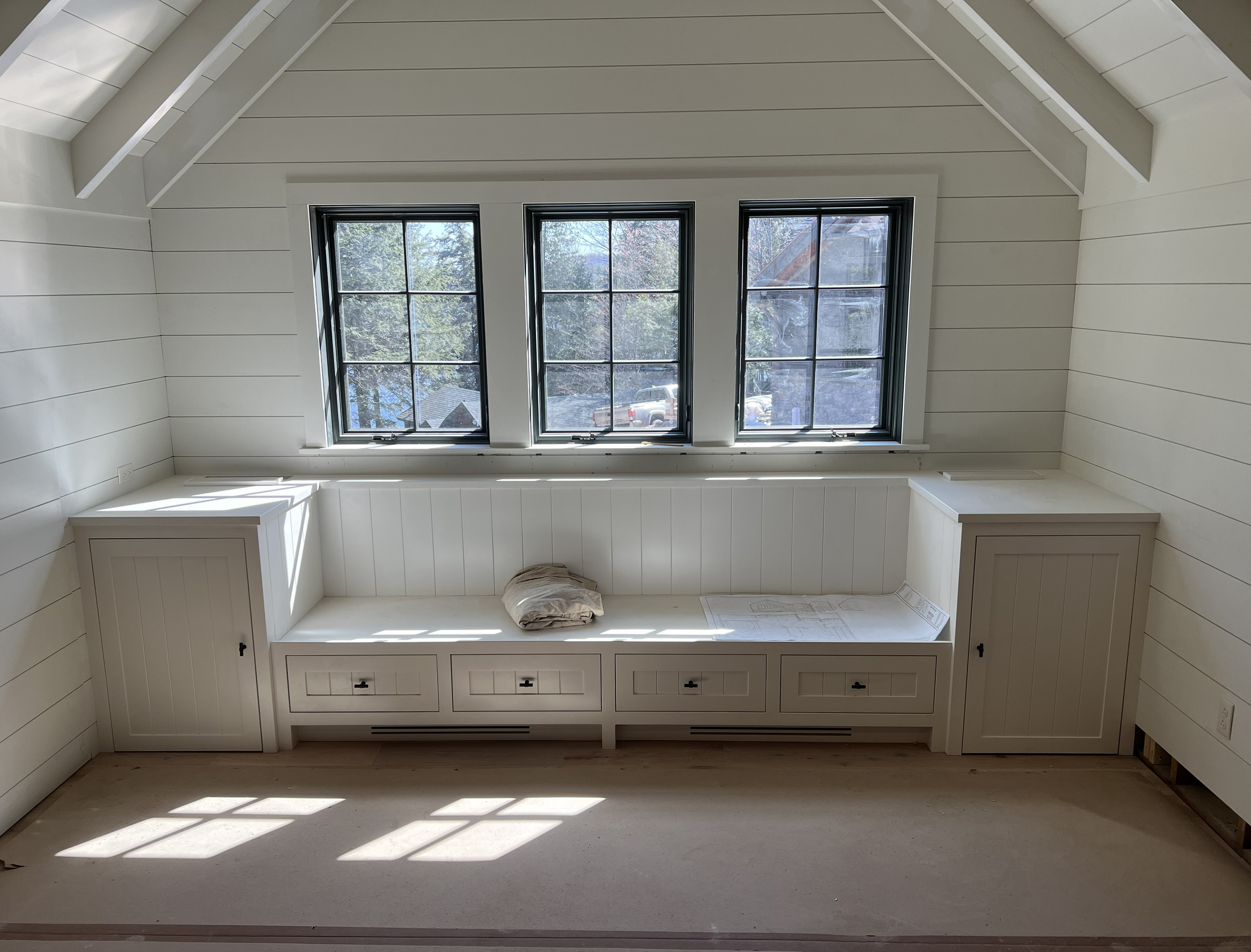 Interior of an attic room with three large windows, white paneled walls, and built-in cabinets below the windows. Construction materials and a stack of rolled-up fabric are on the window bench.