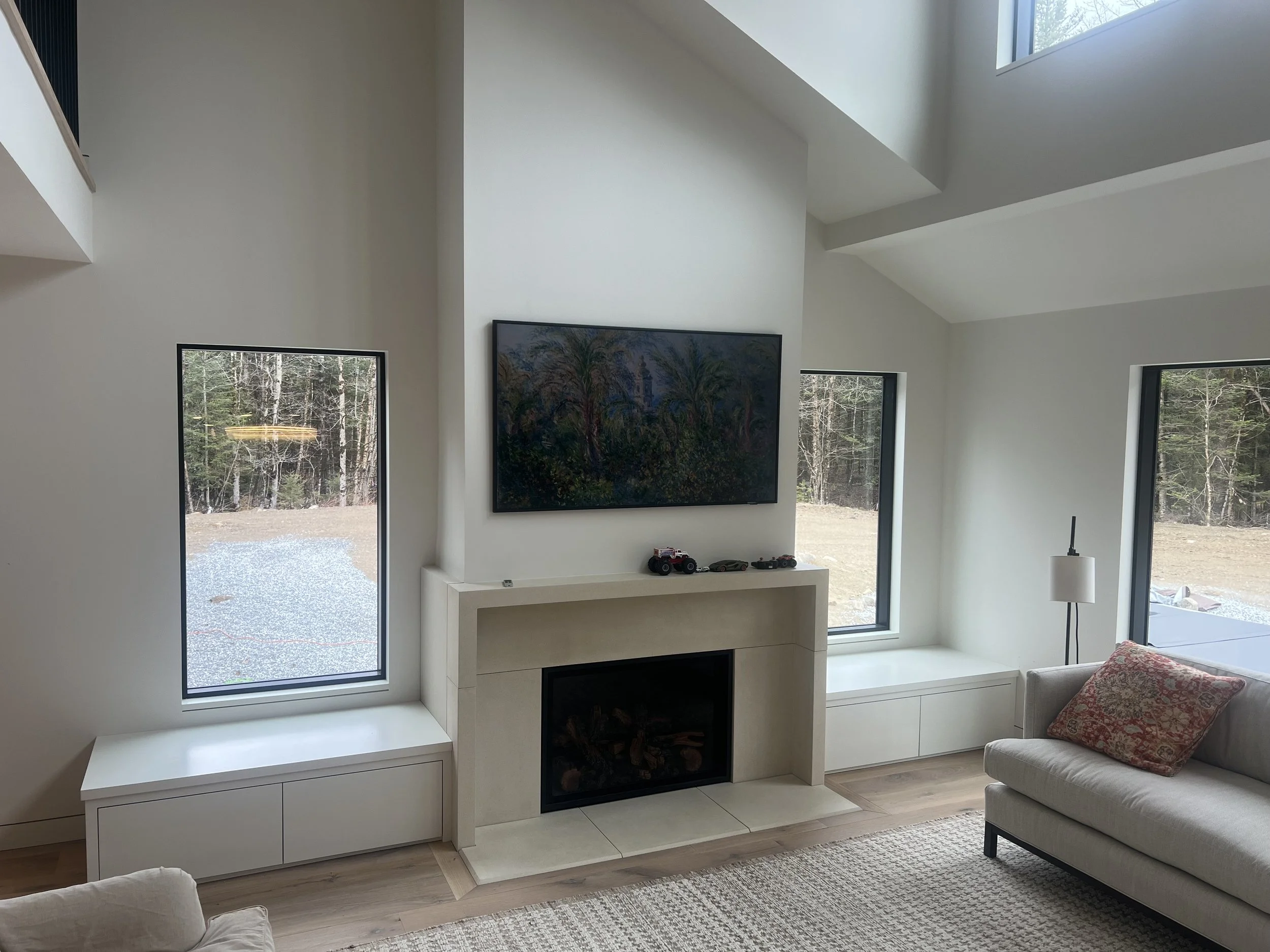 Modern living room with white walls, large windows showing a wooded outdoor scene, a mounted flat-screen TV, a white console with toys below, a cozy armchair with a throw pillow, and a floor lamp.