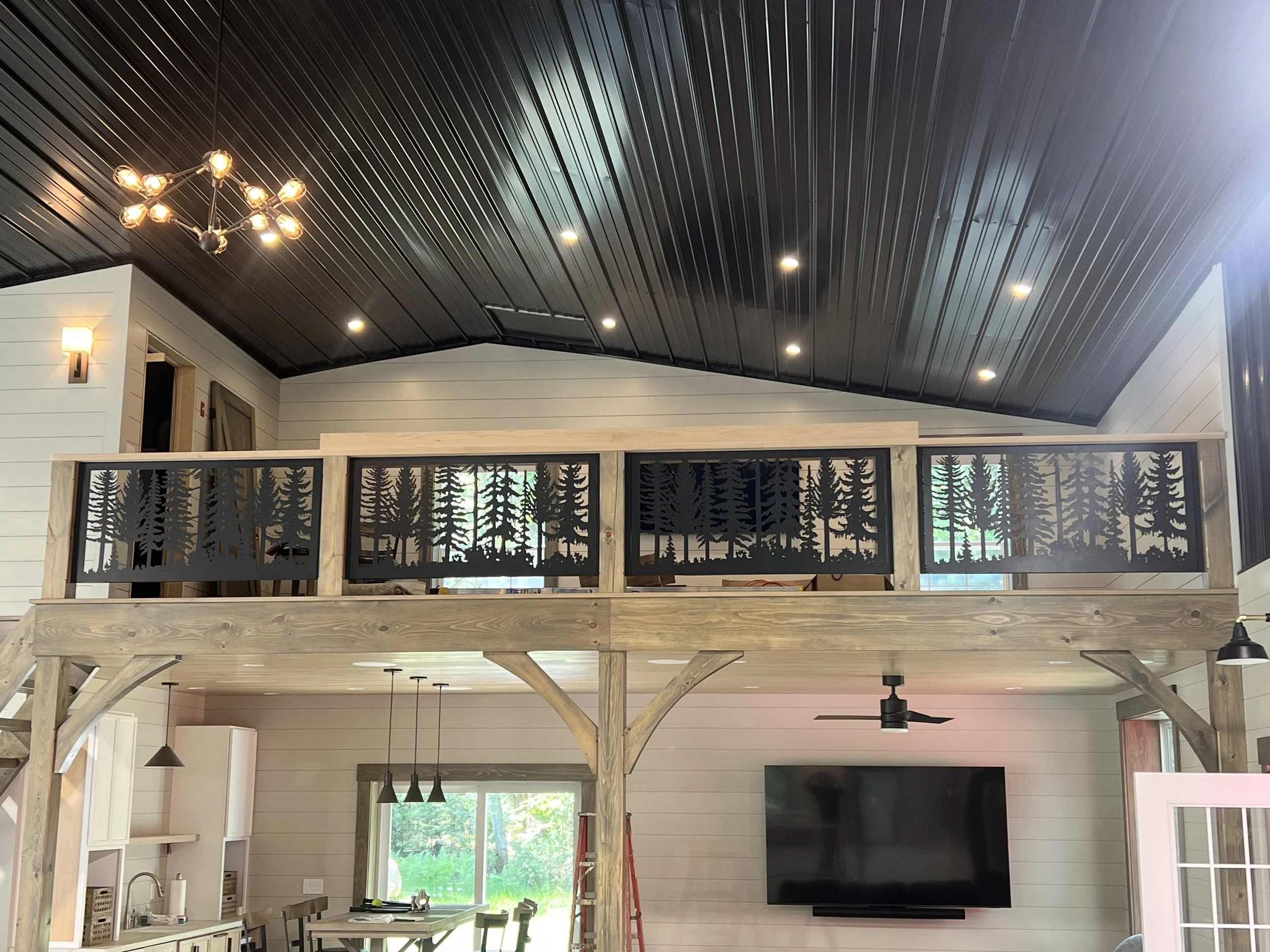 Interior of a house under construction with a high black metal ceiling, wood framing, and modern lighting fixtures, including a chandelier and recessed ceiling lights.