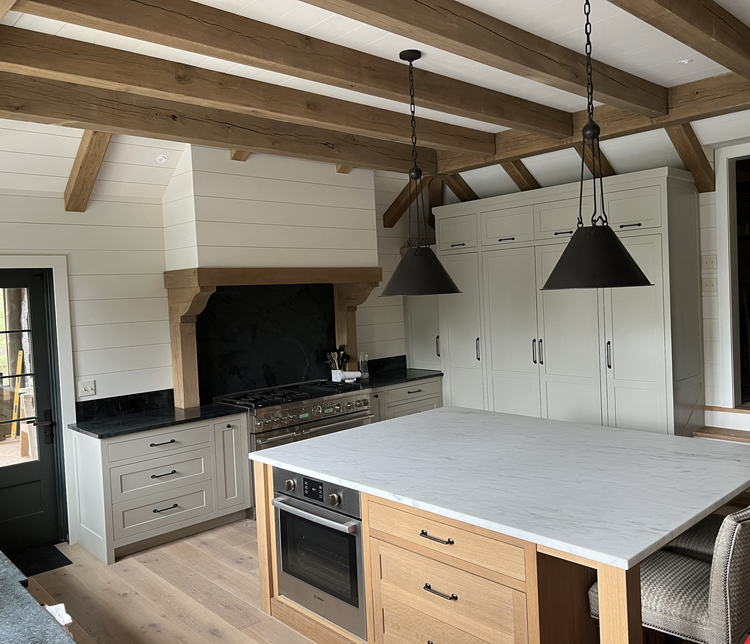 Modern kitchen with white cabinets, black hardware, a gray island with a marble countertop, a stainless steel oven, and wooden ceiling beams with black pendant lights.