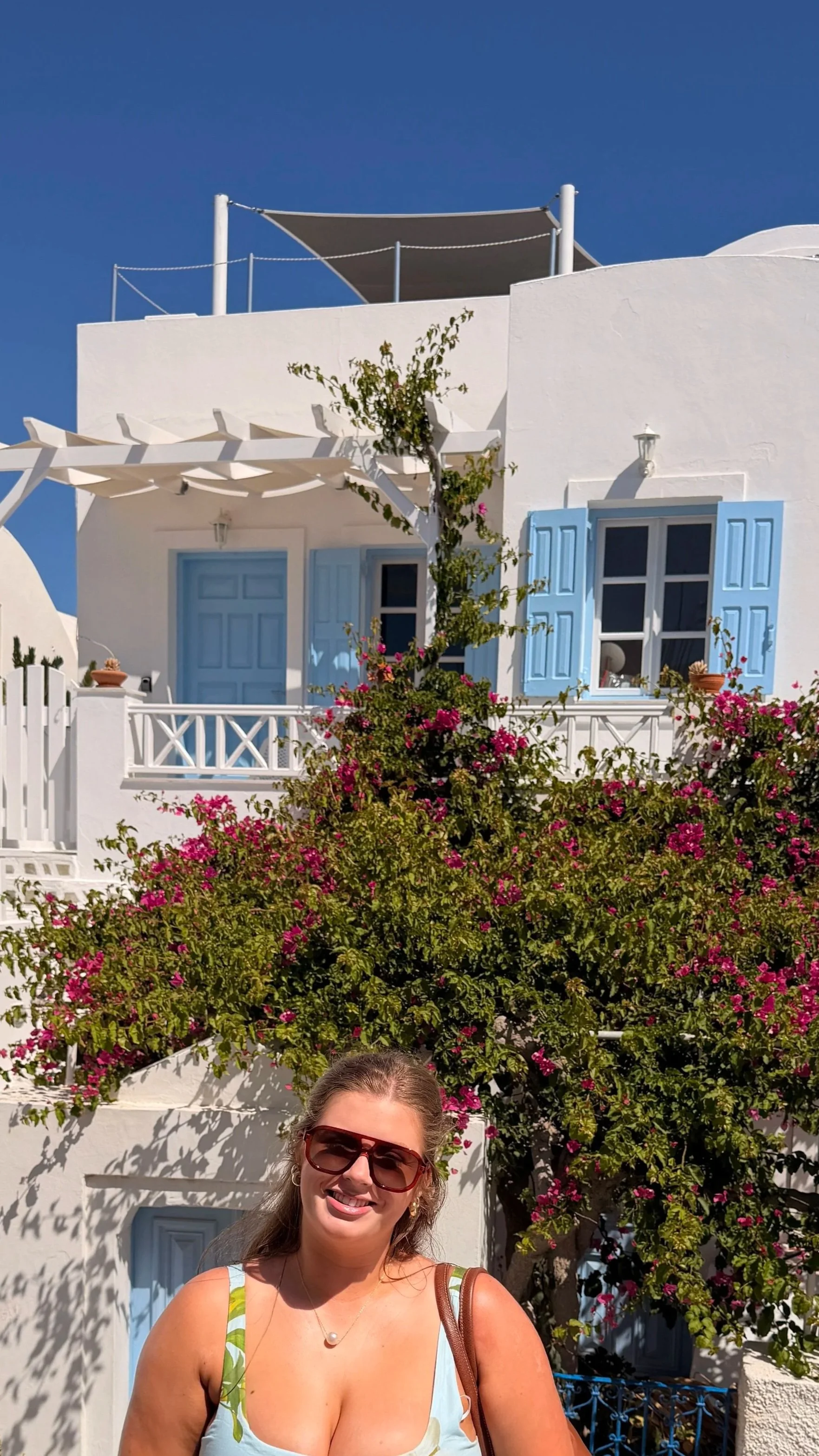 A smiling woman wearing sunglasses and a light-colored dress with green and yellow patterns, standing in front of a white building with blue accents, pink flowers, and greenery.