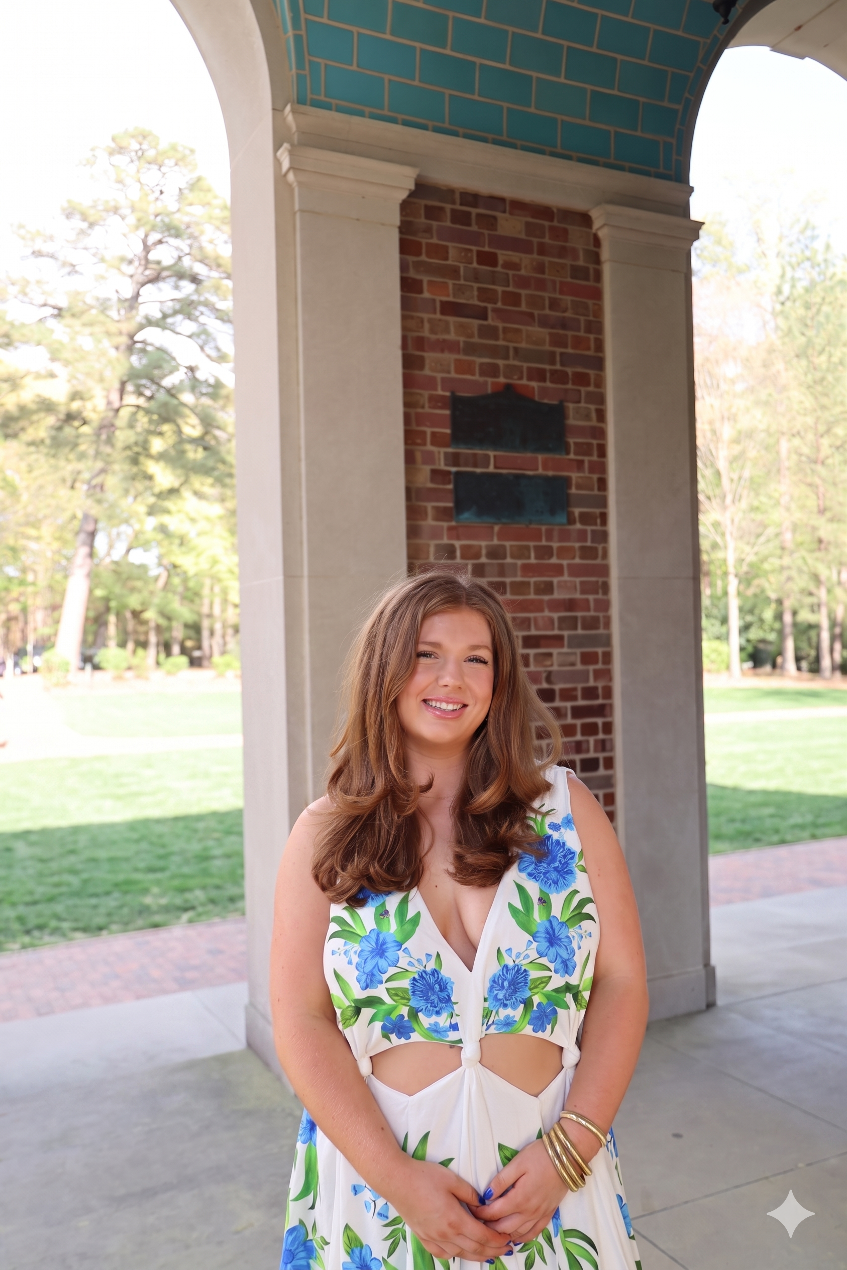 A young woman with long, wavy brown hair smiling in front of a brick and stone pavilion, wearing a white dress with large blue flowers and green leaves, with her hands clasped in front of her.