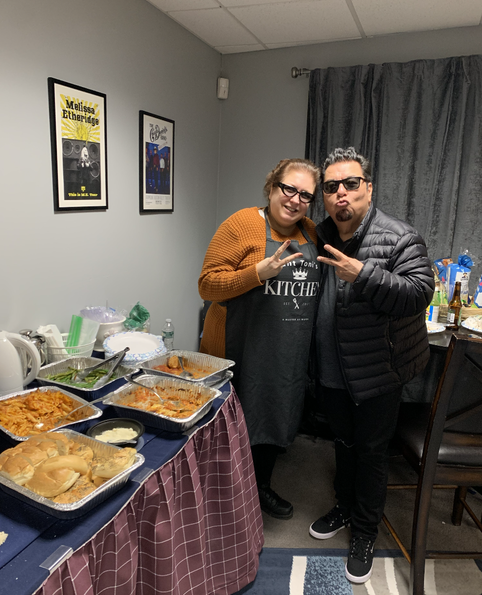 Two people wearing sunglasses posing together at a party, standing next to a table with food, including bread rolls, pasta, and other dishes, in a room with framed posters on the wall.