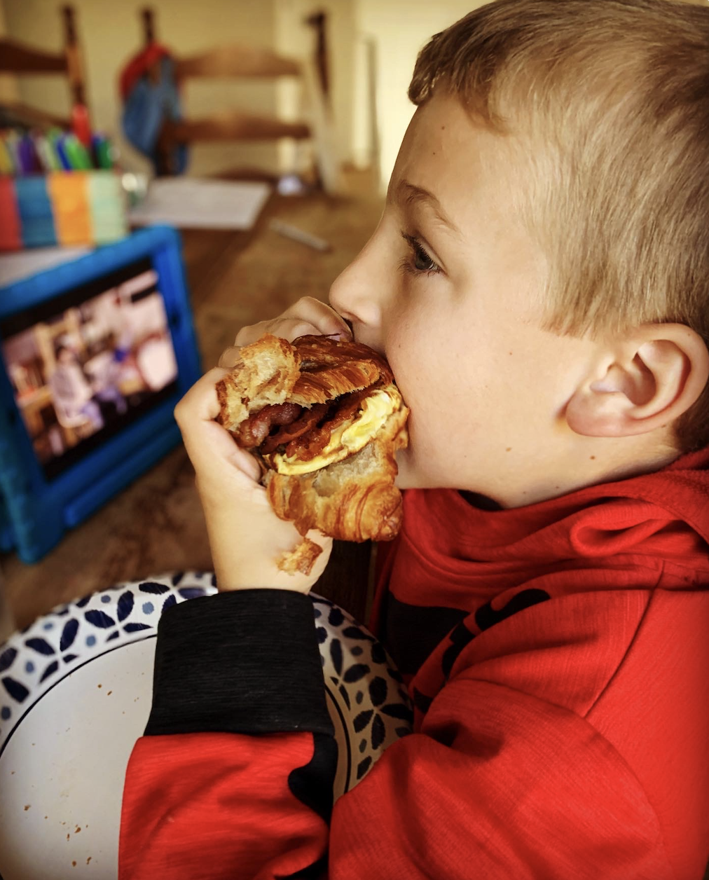 A young boy in a red hoodie eating a sandwich with bacon and scrambled eggs, sitting at a table with a patterned plate. In the background, a blue TV, books, and a wooden chair are visible.