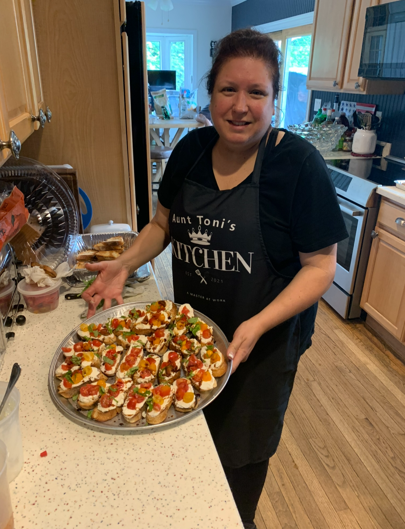 A woman wearing a black apron with the words "Aunt Toni's Kitchen" is holding a large tray of colorful bruschetta topped with various chopped vegetables and herbs in a home kitchen.