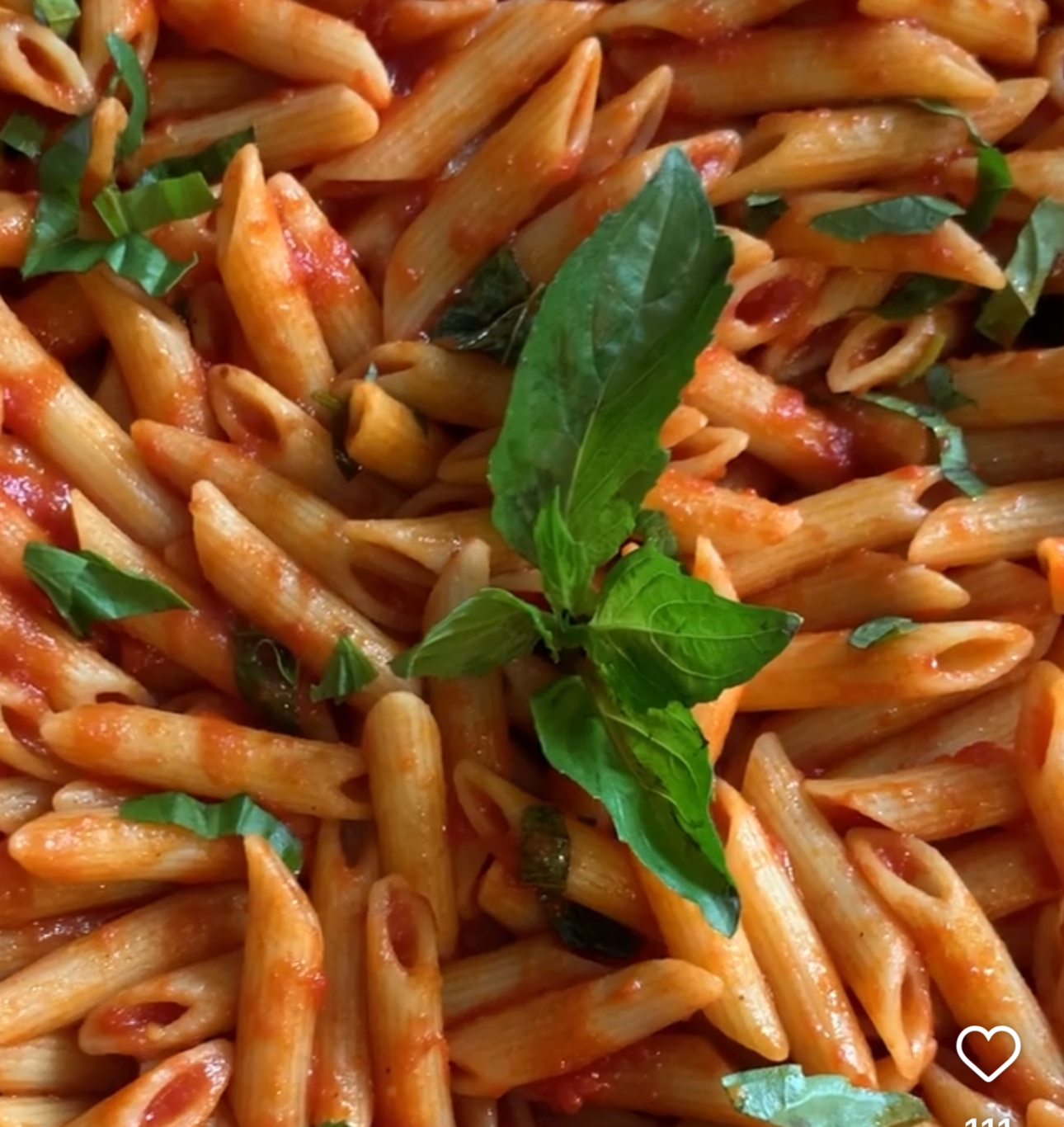 Close-up of cooked penne pasta with tomato sauce, garnished with fresh basil leaves and chopped herbs.