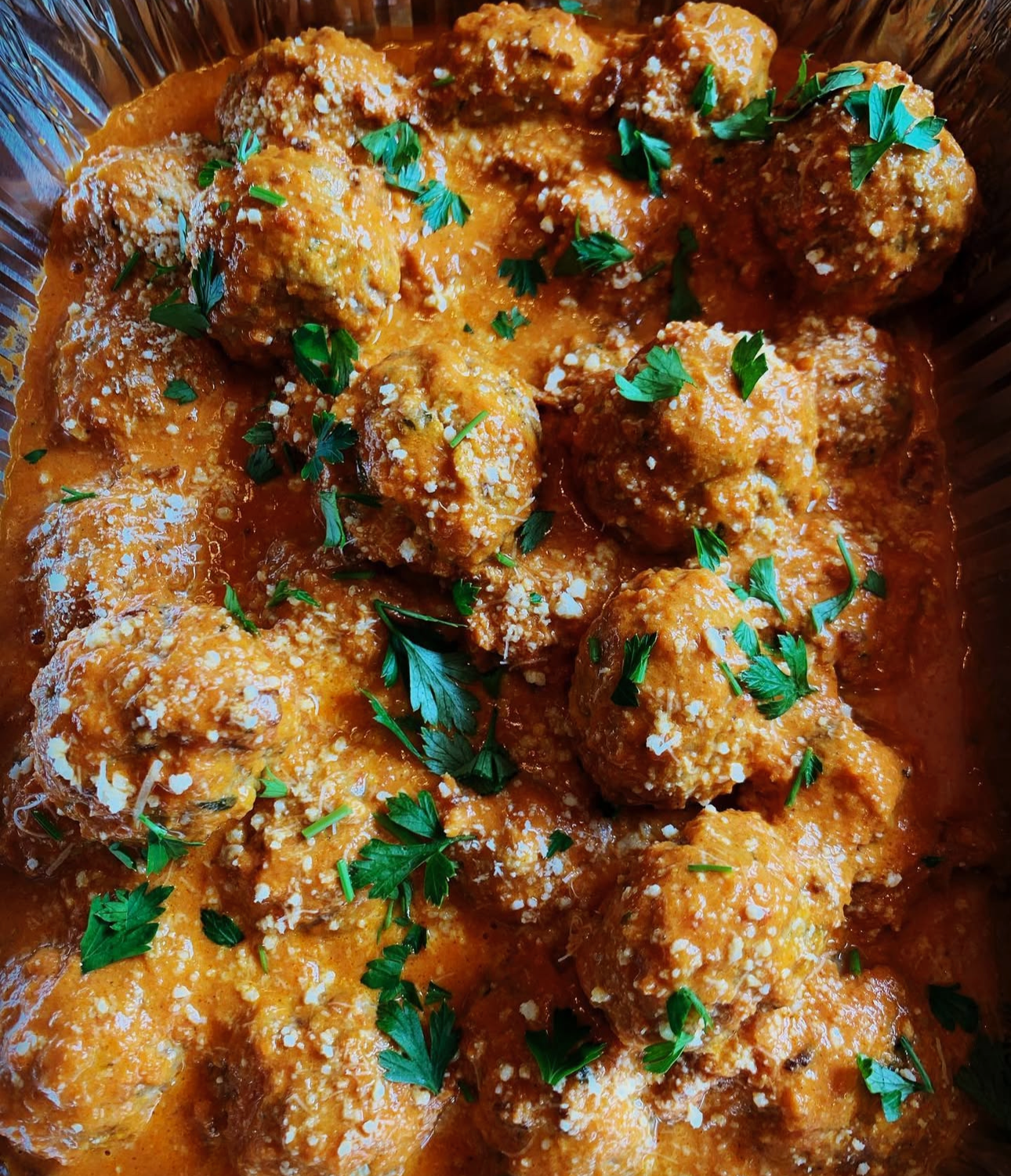 Plate of cooked meatballs covered in tomato sauce, garnished with chopped parsley and grated cheese.