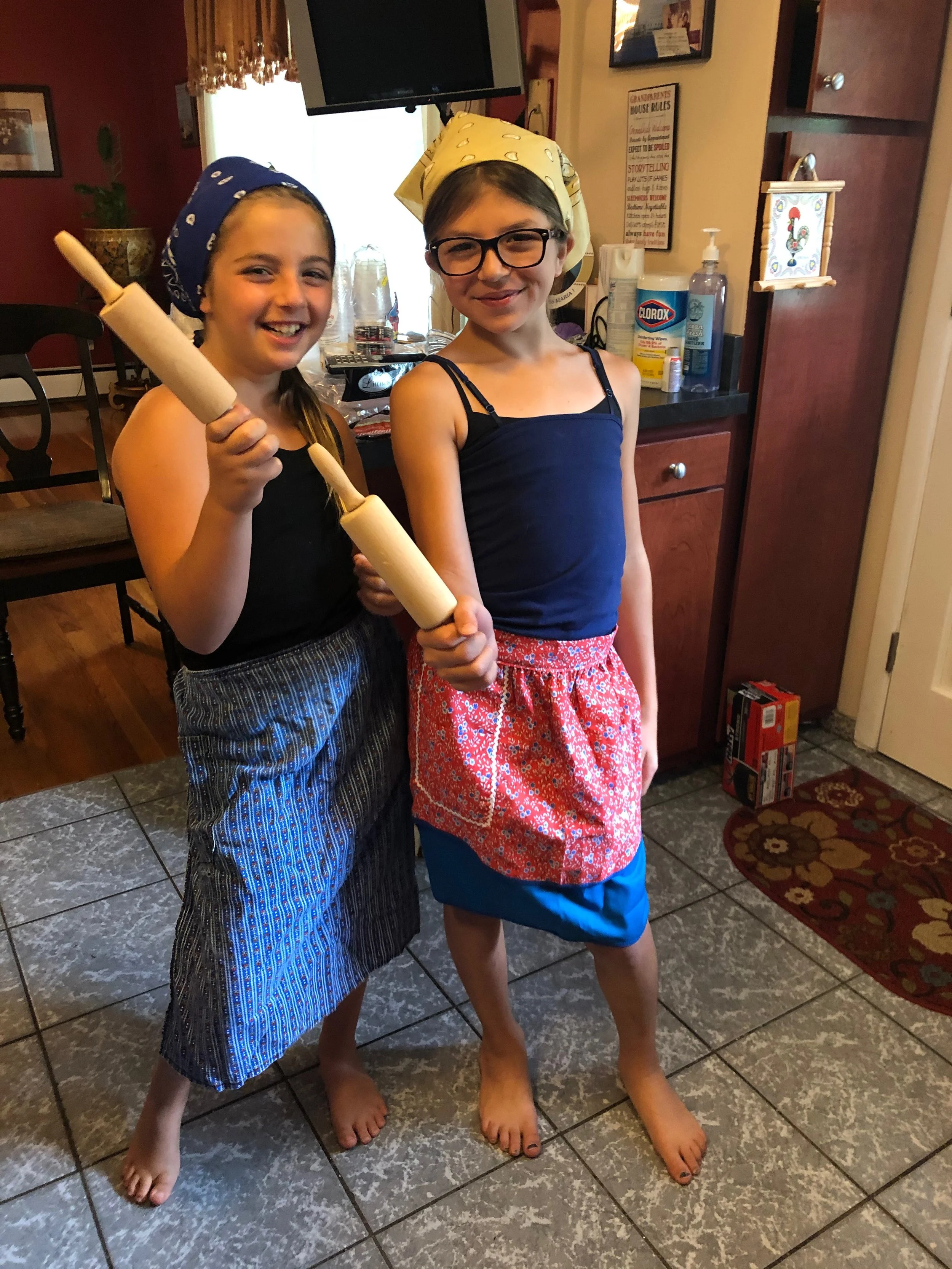 Two girls standing in a kitchen, smiling and holding rolling pins with bandanas on their heads, one in a black top and striped shorts, the other in a blue dress with a red patterned apron over blue shorts.