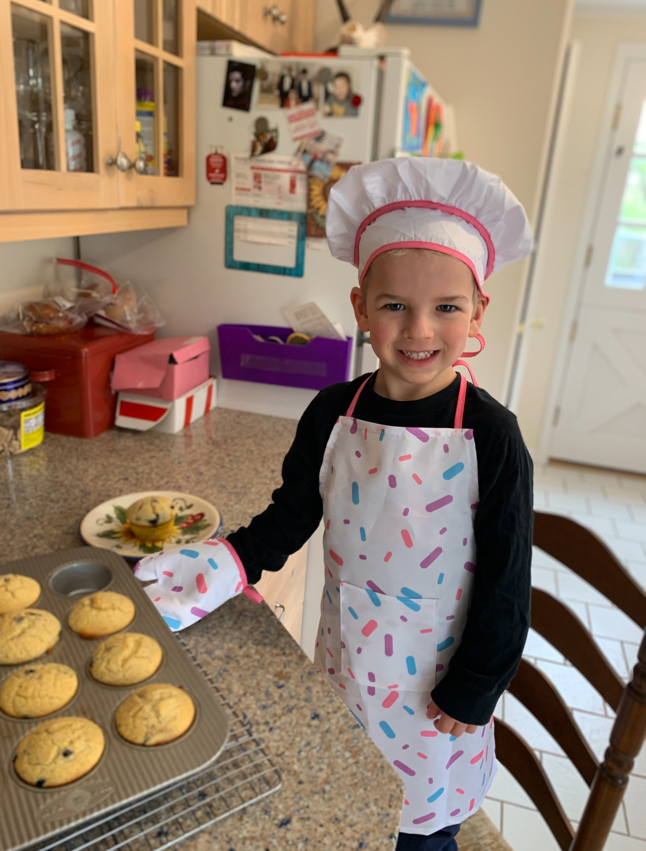 Young boy wearing a white chef's hat and apron with colorful sprinkles pattern, smiling while baking cookies in a kitchen.