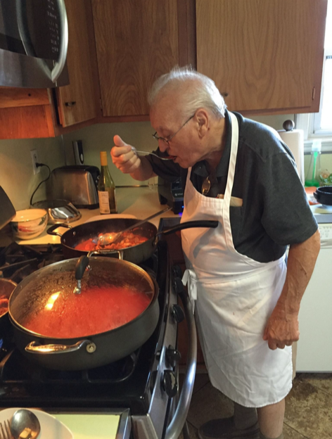 An elderly man cooking on a stove wearing a white apron, tasting the sauce with a spoon, in a kitchen with wooden cabinets and various kitchen items.