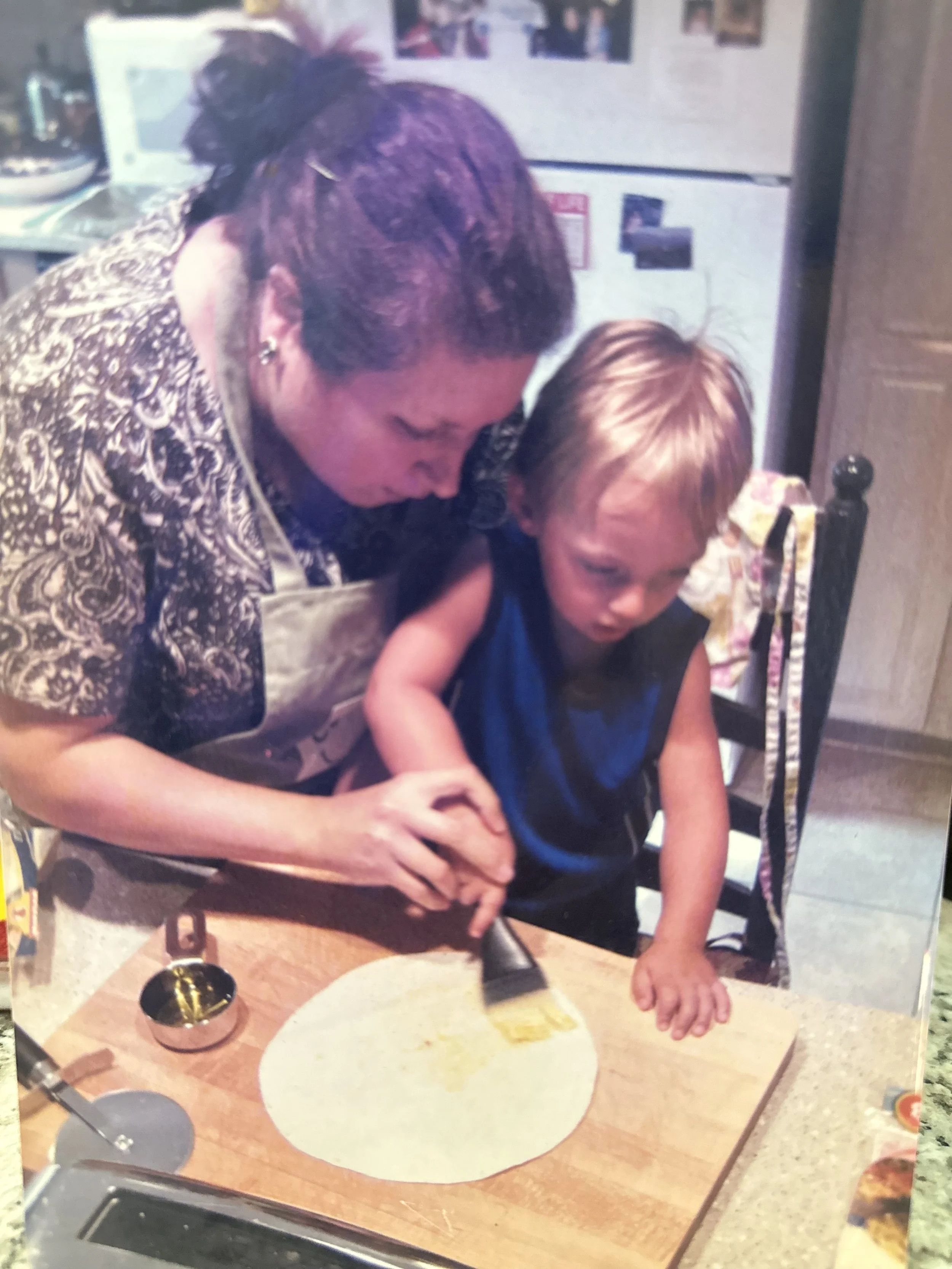 A woman with short hair leaning over a girl with red hair, who is sitting at a kitchen table with a large plate of food. The woman appears to be helping or assisting the girl. The background includes a refrigerator with photos and magnets, and a kitchen counter with various items.