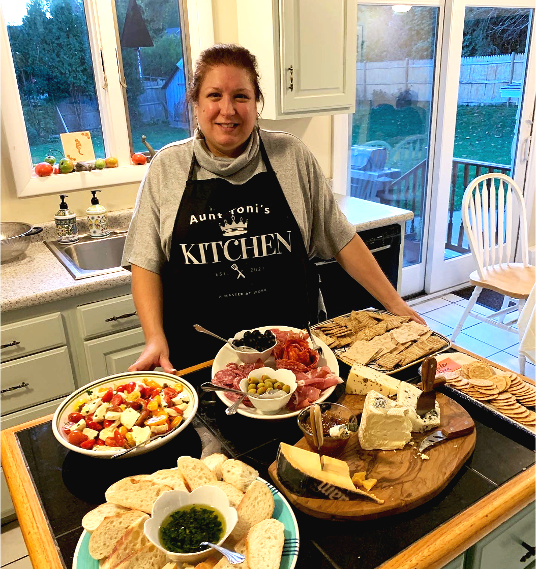 A woman smiling in a kitchen, wearing a black apron that says 'Aunt Toni's Kitchen.' There are various cheeses, crackers, a vegetable salad, bread, cured meats, slices of cheese, and condiments on the counter in front of her, with a window and dining area in the background.