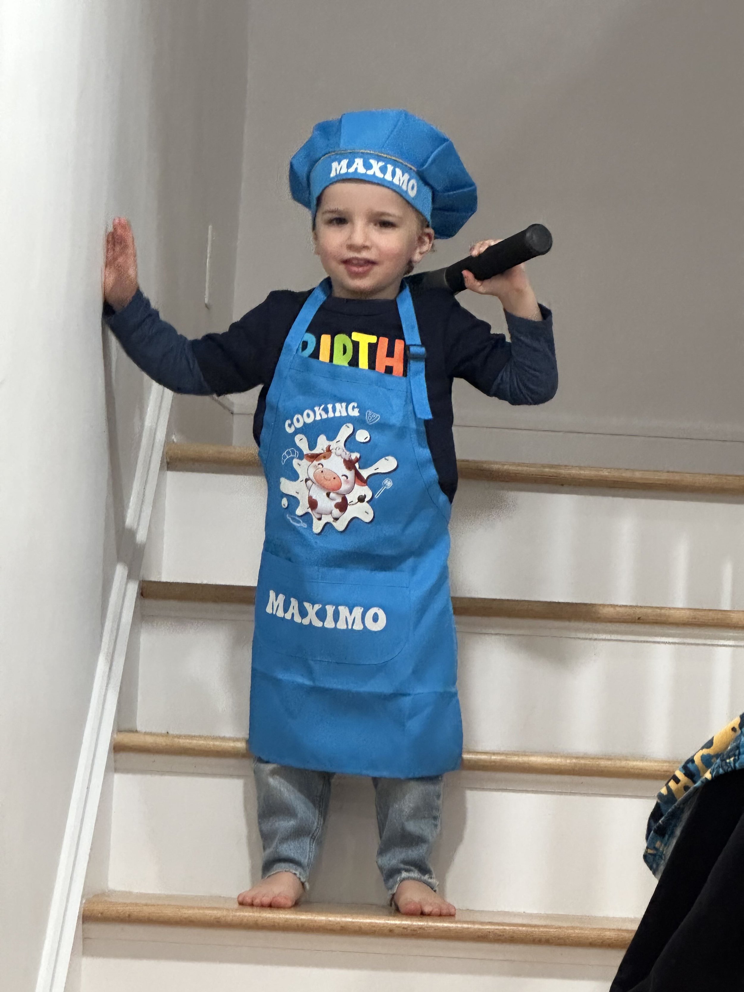 Young boy standing on stairs wearing a blue chef's hat and apron, holding a black toy sword, with a colorful birthday shirt and gray pants.