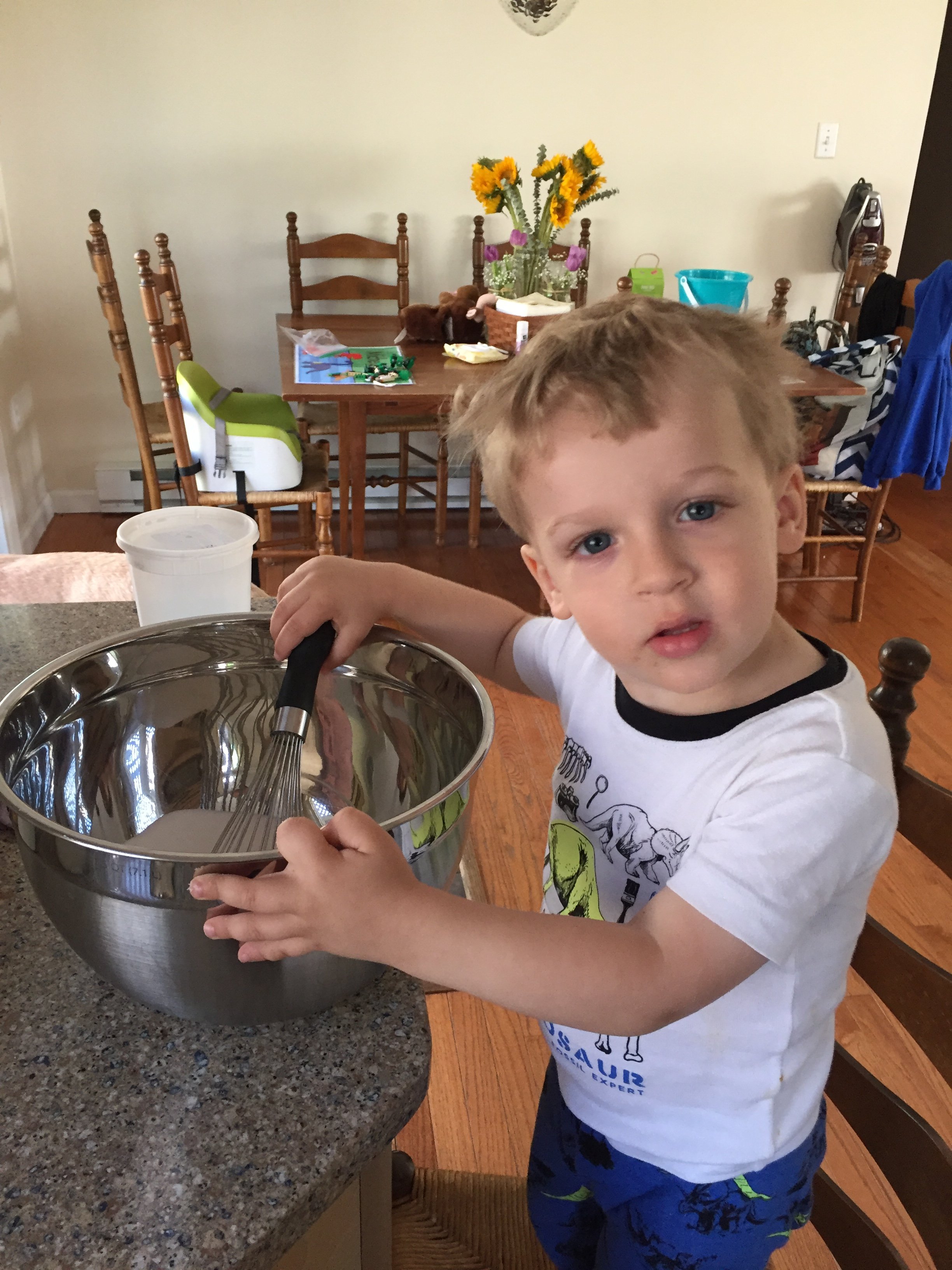 Young boy with blonde hair and blue eyes stirring a large stainless steel mixing bowl in a kitchen, wearing a white graphic t-shirt and blue shorts with green dinosaur patterns.