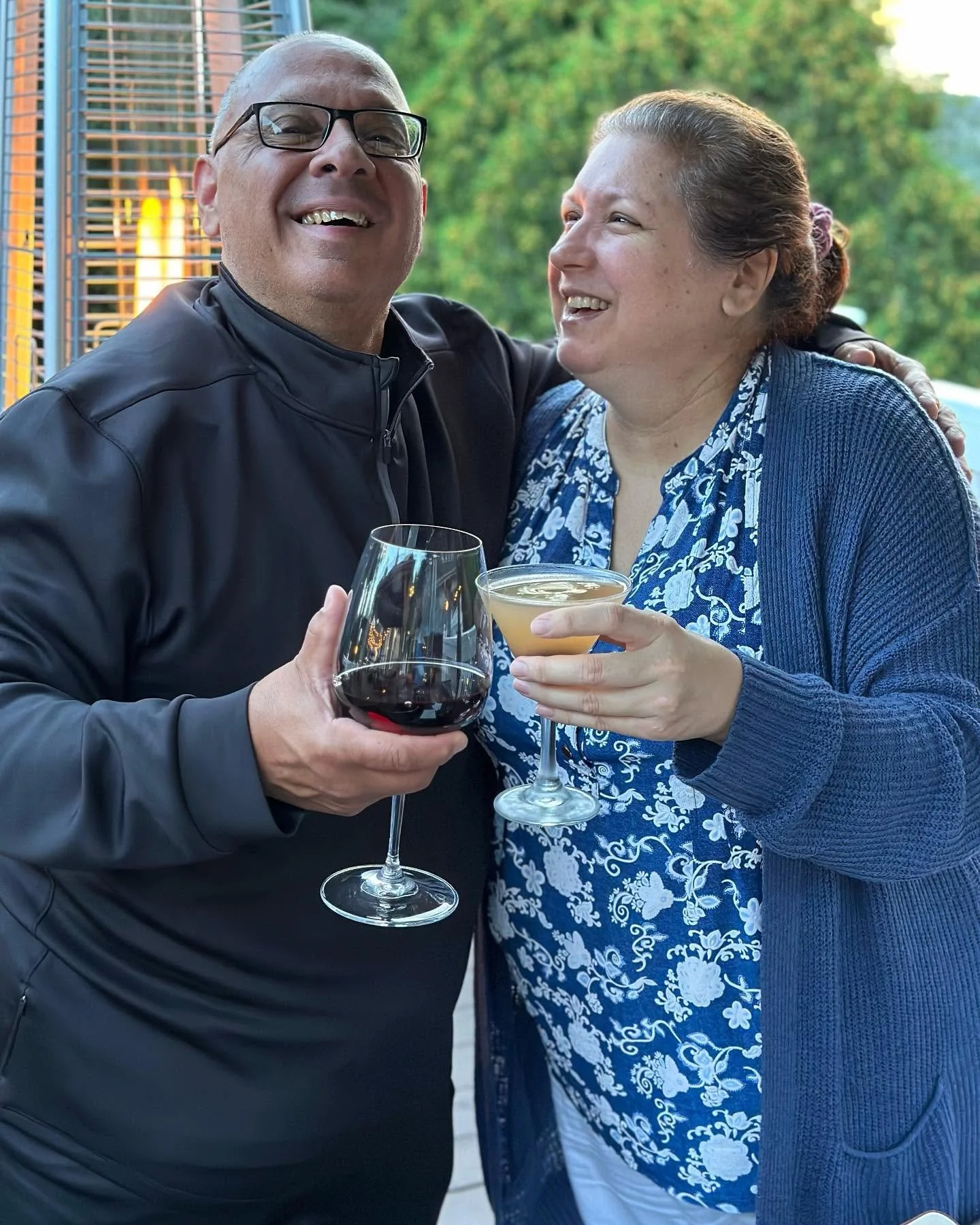 Man and woman smiling, clinking glasses of wine and cocktail outdoors with green trees in the background.