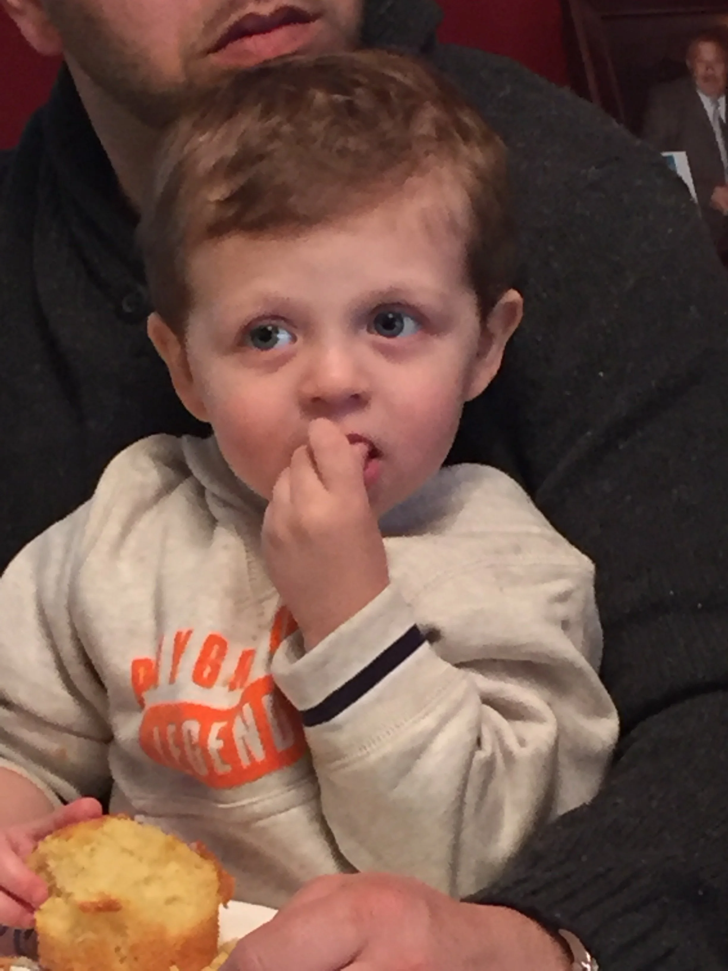 A young boy with red hair and light skin eating a slice of yellow cake, while sitting on an adult's lap indoors.