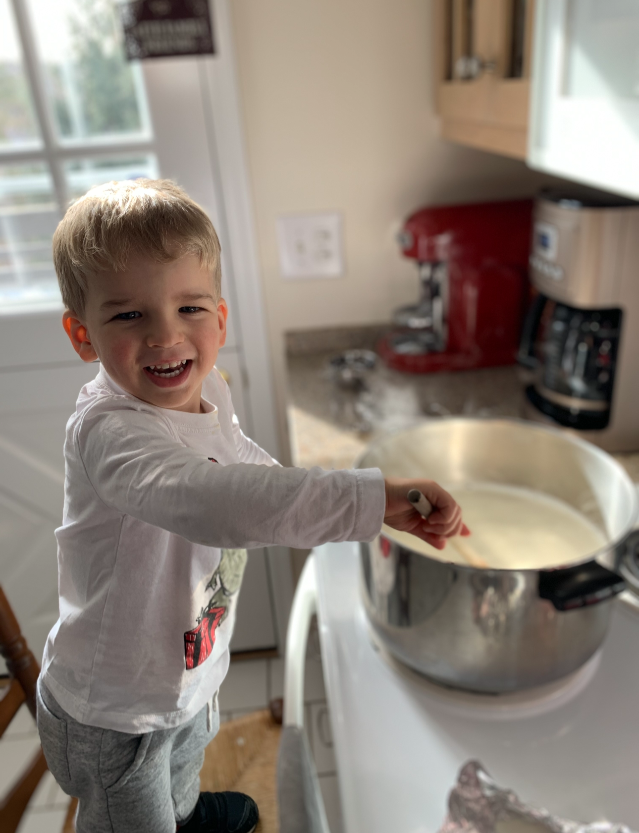 A young boy with blond hair and a white long-sleeve shirt is smiling and stirring a large mixing bowl in a kitchen.
