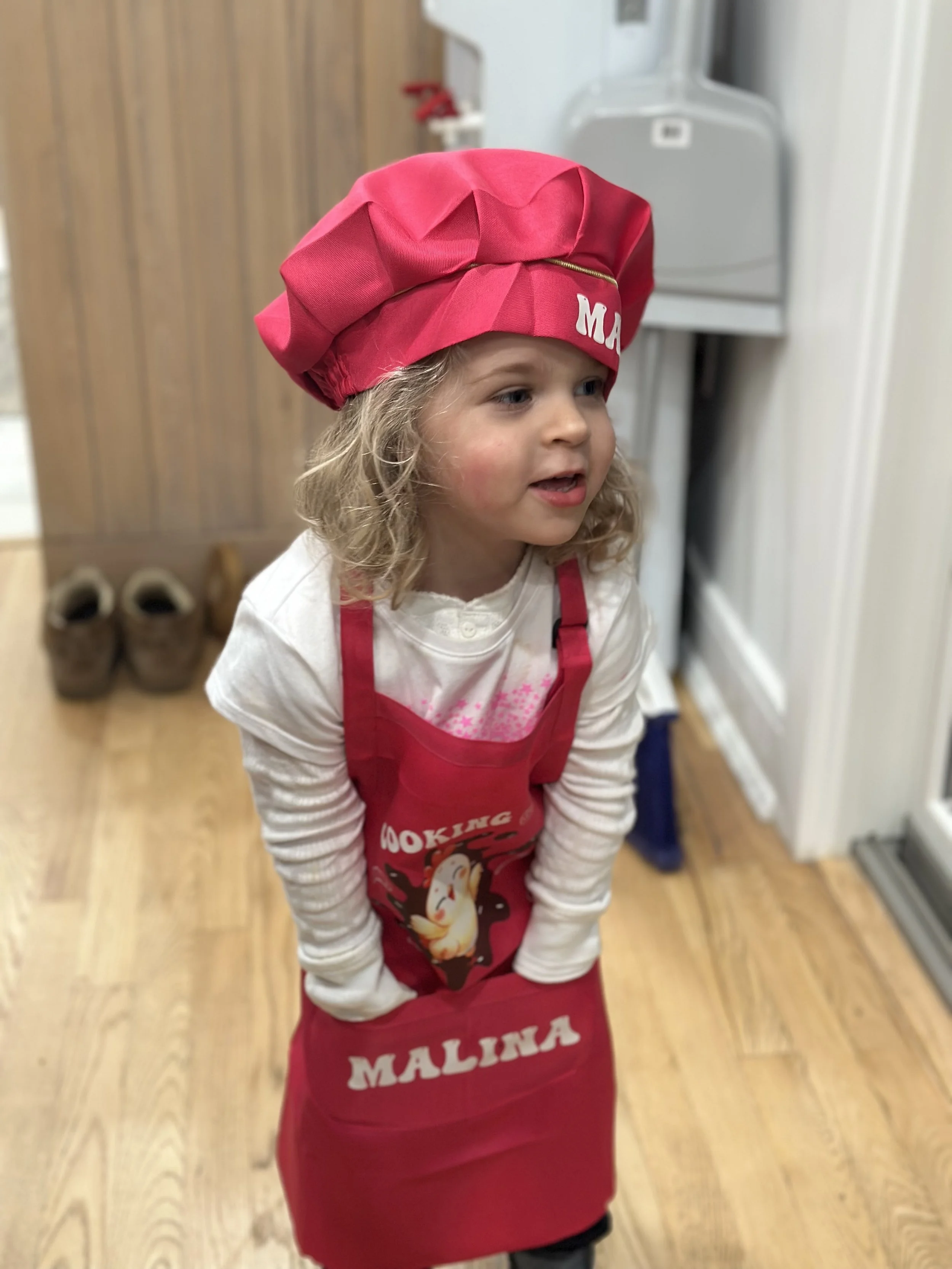 A young girl wearing a red chef's hat and apron with the word 'MALINA' on it, standing indoors on a wooden floor.