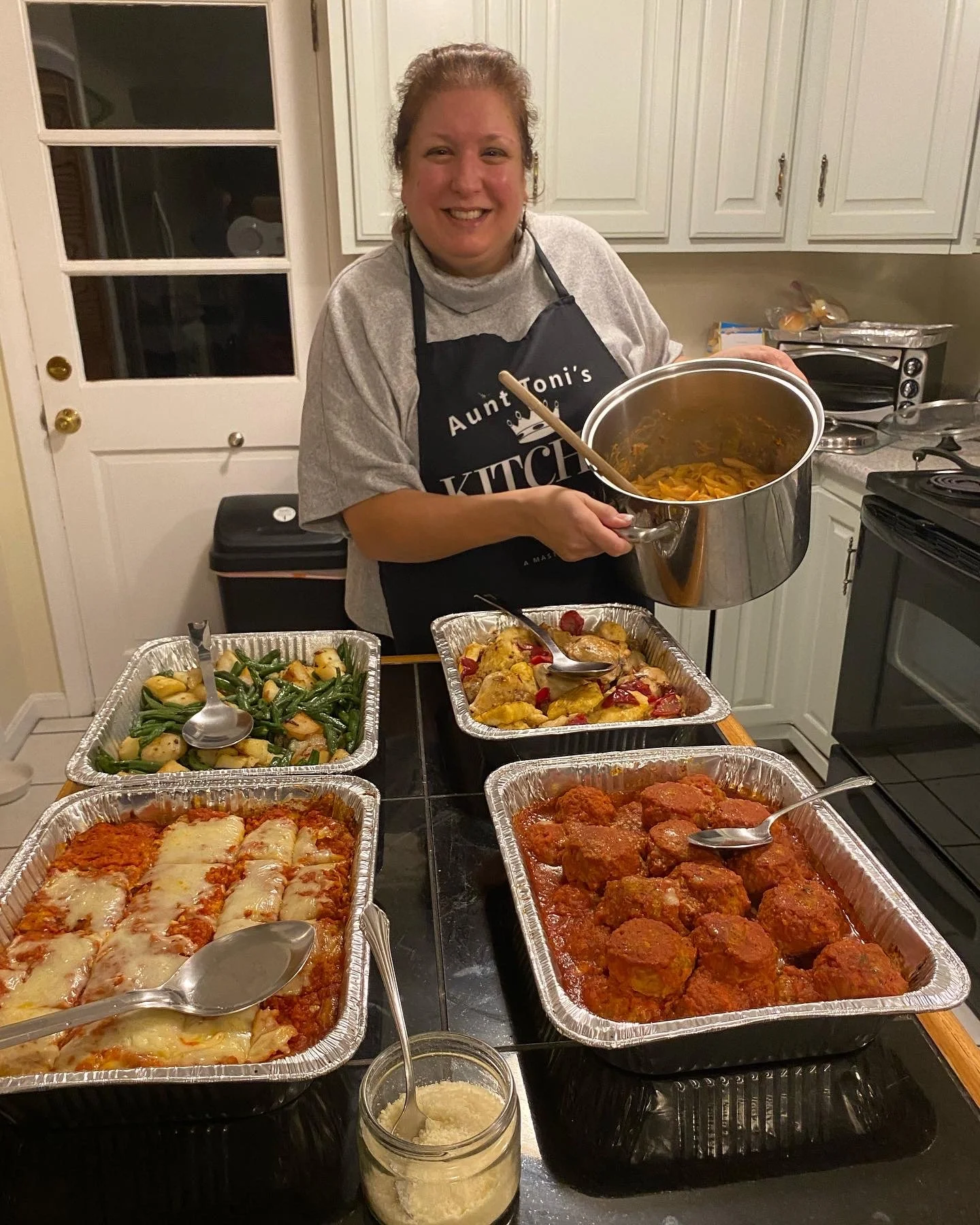 A woman smiling in a kitchen, holding a pot of cooked pasta, with several trays of cooked Italian dishes on the counter including meatballs with tomato sauce, baked lasagna, and chicken with vegetables.