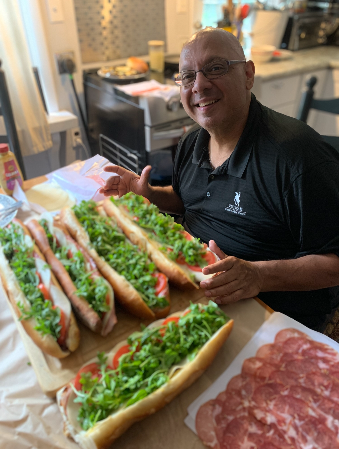 A smiling man wearing glasses and a black polo shirt is holding a tray of sandwich subs loaded with tomatoes, lettuce, and green herbs. The kitchen background is blurred, with kitchen appliances and items visible on the counter.