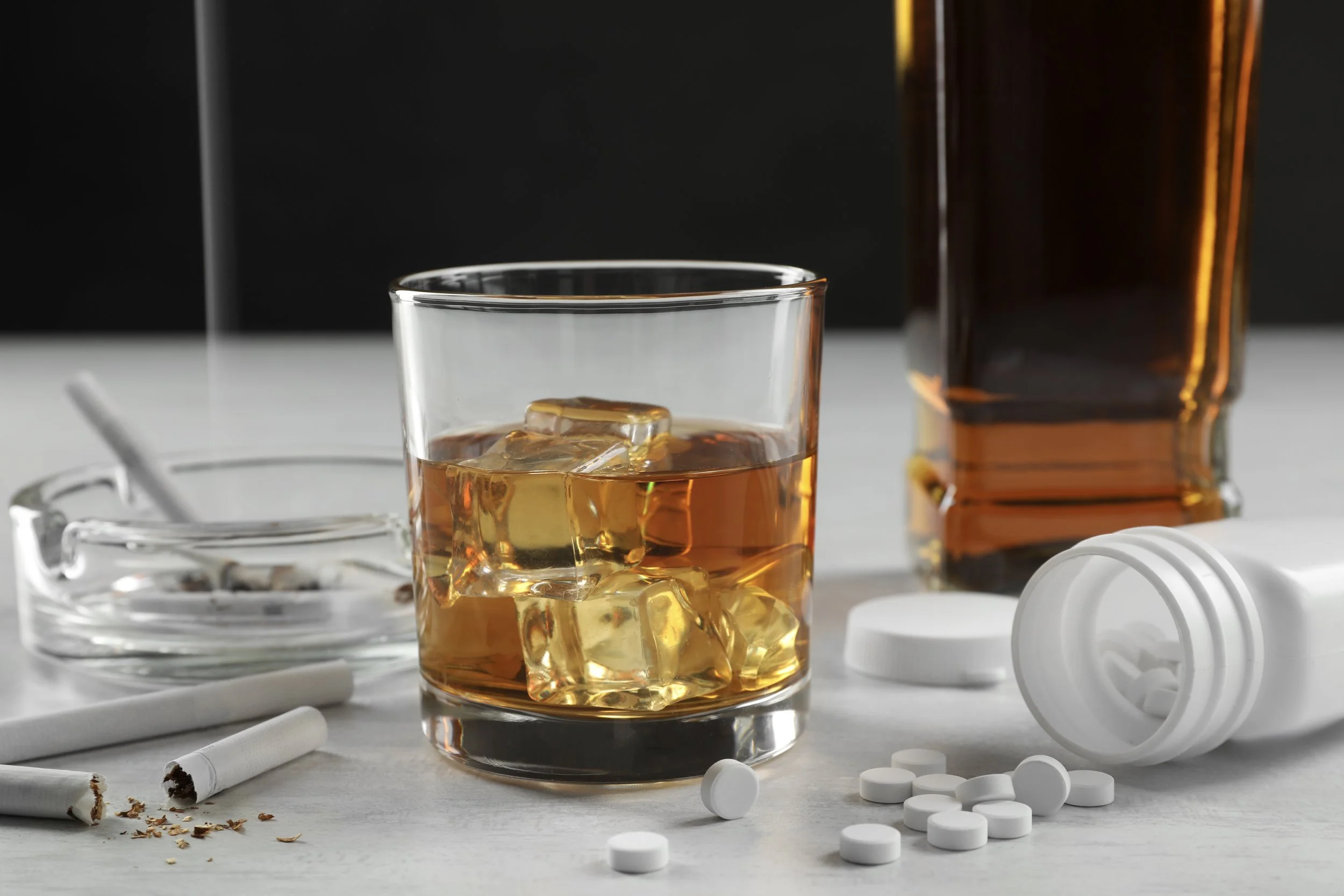 A glass of amber liquor with ice, surrounded by cigarette butts, scattered white pills, and an orange pill bottle on a white surface.