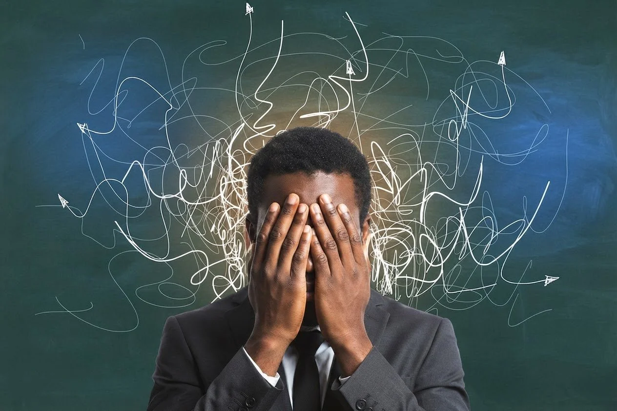 Man wearing a suit with hands covering his face, against a chalkboard background with chaotic white lines and arrows.