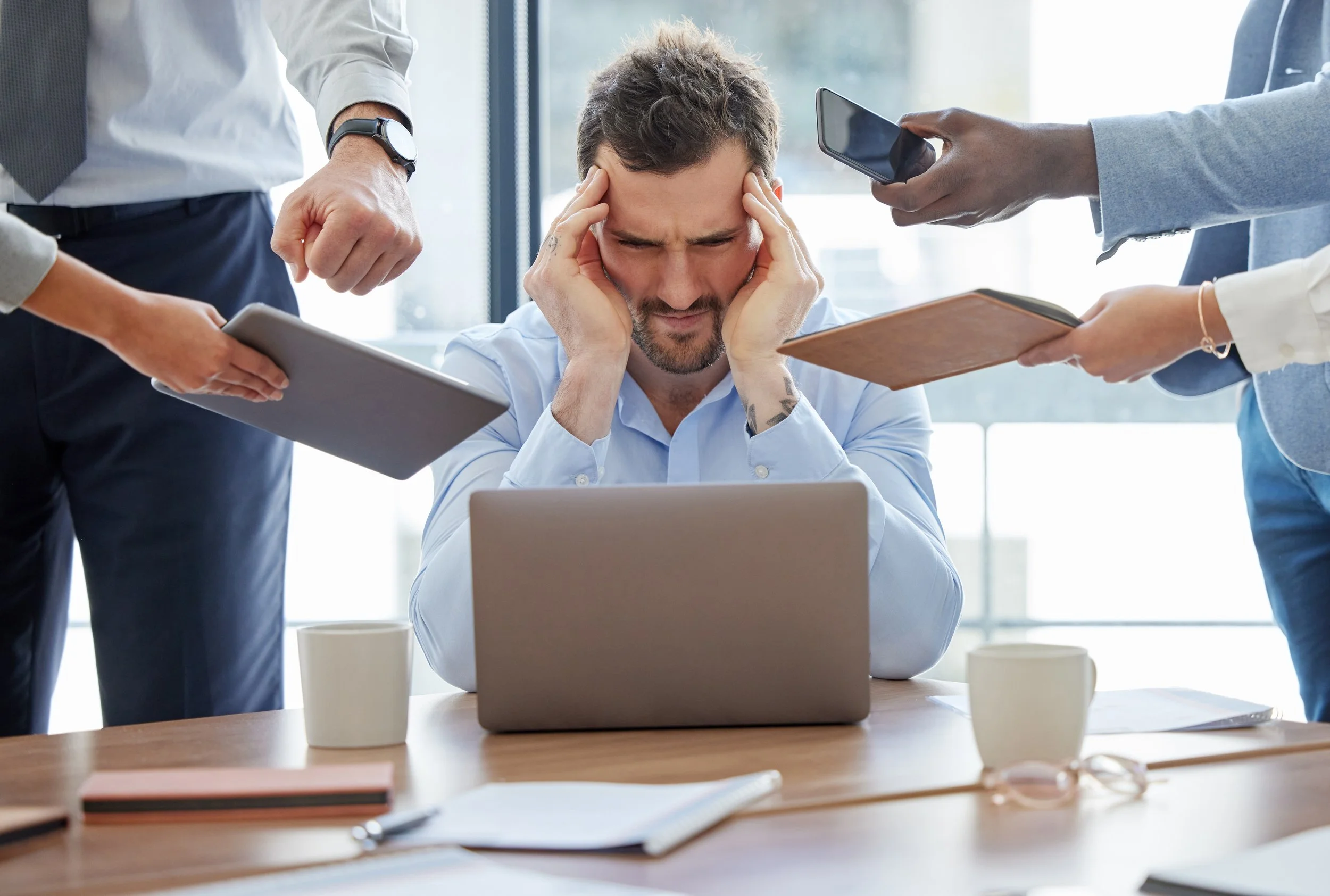 A man sitting at a table with his head in his hands, appearing stressed while surrounded by four people pointing at him and taking notes, in a modern office setting.