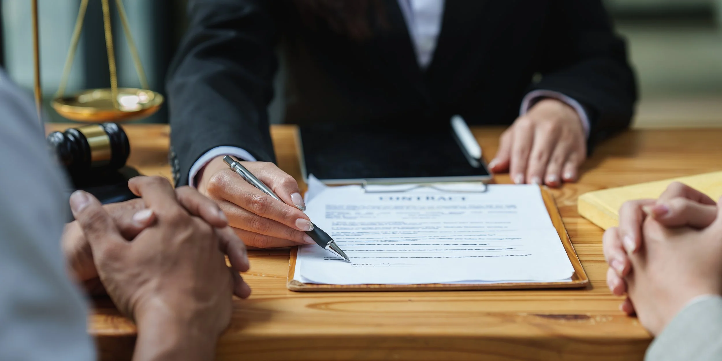 Someone signing a legal document during a meeting with multiple people. A lawyer's gavel and scales of justice are visible in the background.
