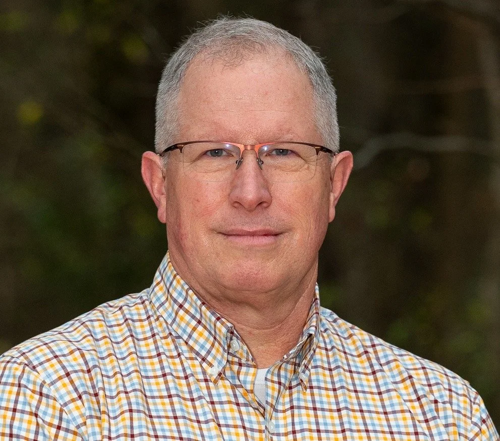 Headshot of a middle-aged man with short gray hair, glasses, and a checkered button-up shirt, outdoors with blurred greenery background.