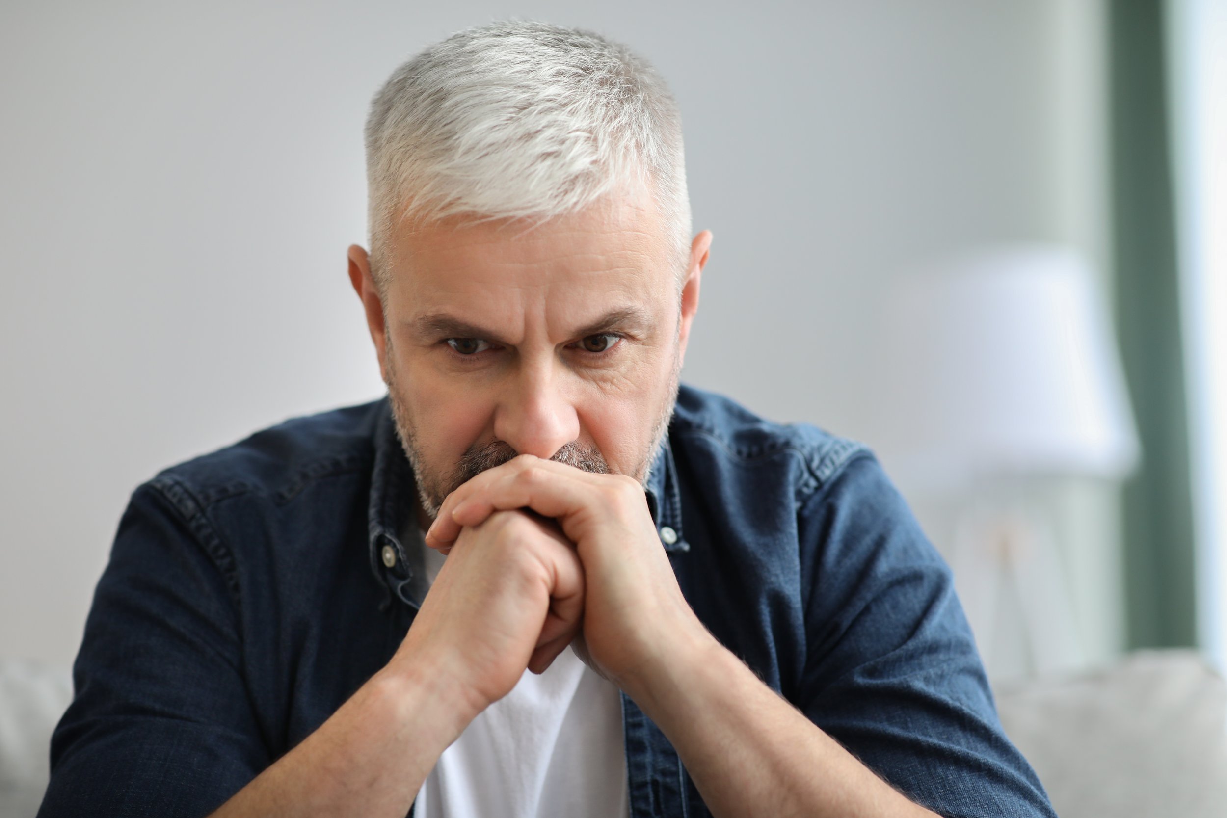 A middle-aged man with gray hair and a beard resting his chin on his hands, deep in thought, wearing a dark denim shirt over a white T-shirt, sitting in a softly lit room.