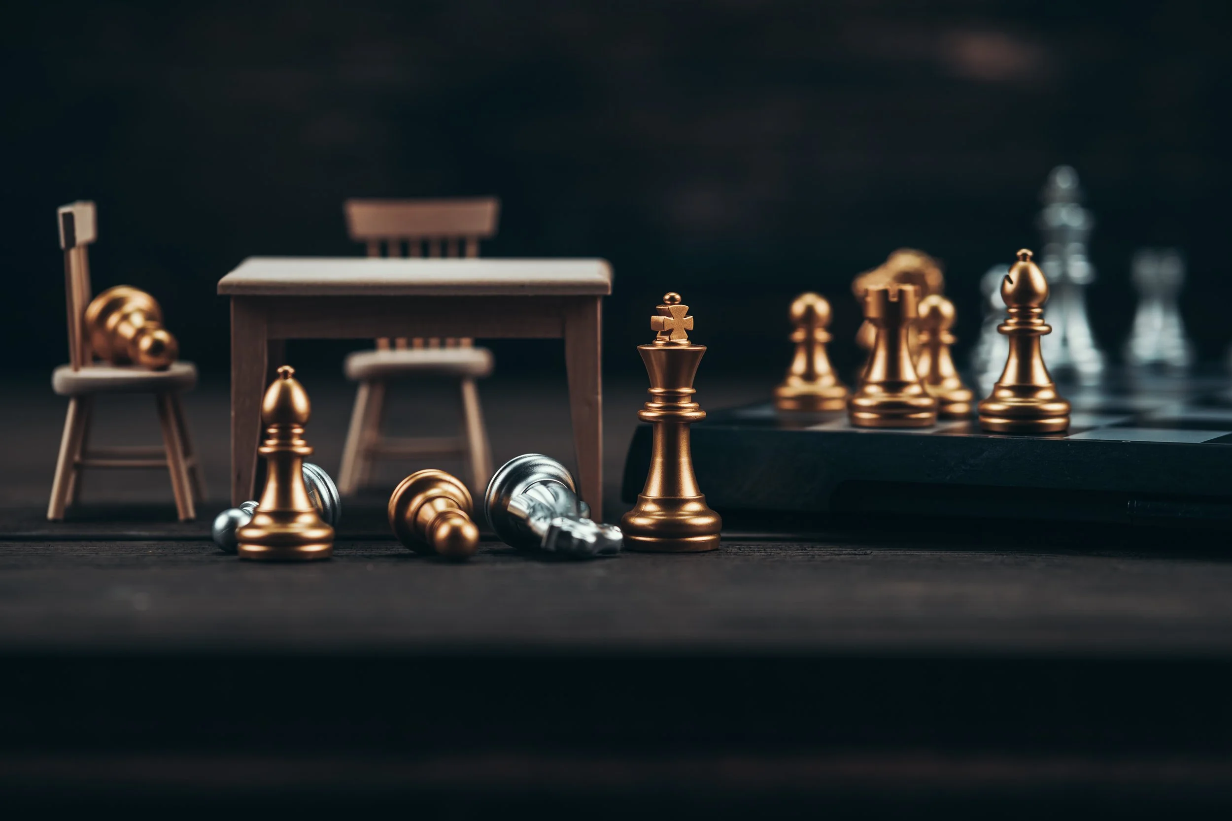 Close-up of metallic chess pieces on a chessboard, with some pieces knocked over, with a small wooden desk and chair in the background.