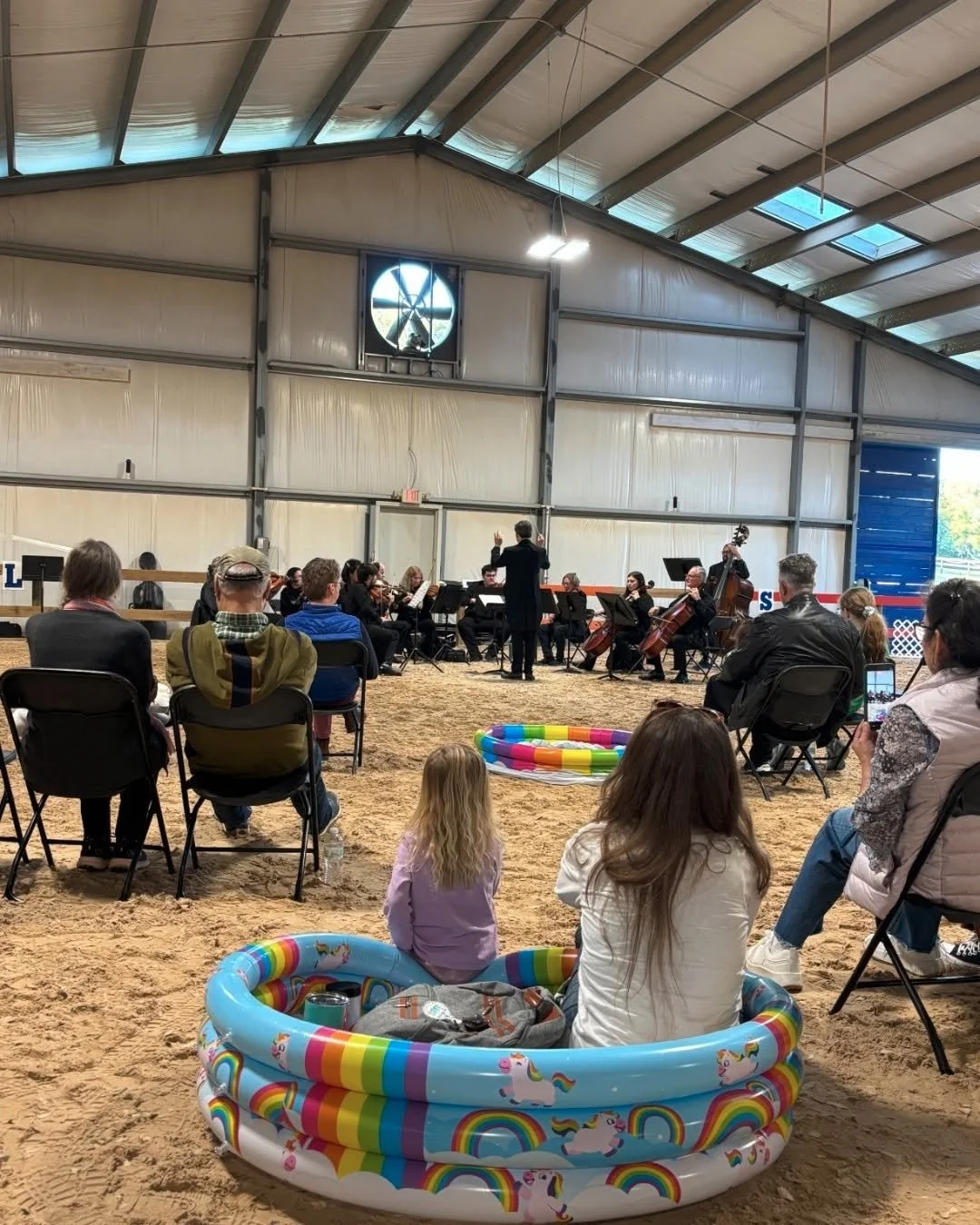 An indoor concert of a string orchestra with a conductor, audience seated on chairs, and children sitting in a rainbow unicorn-themed inflatable pool.