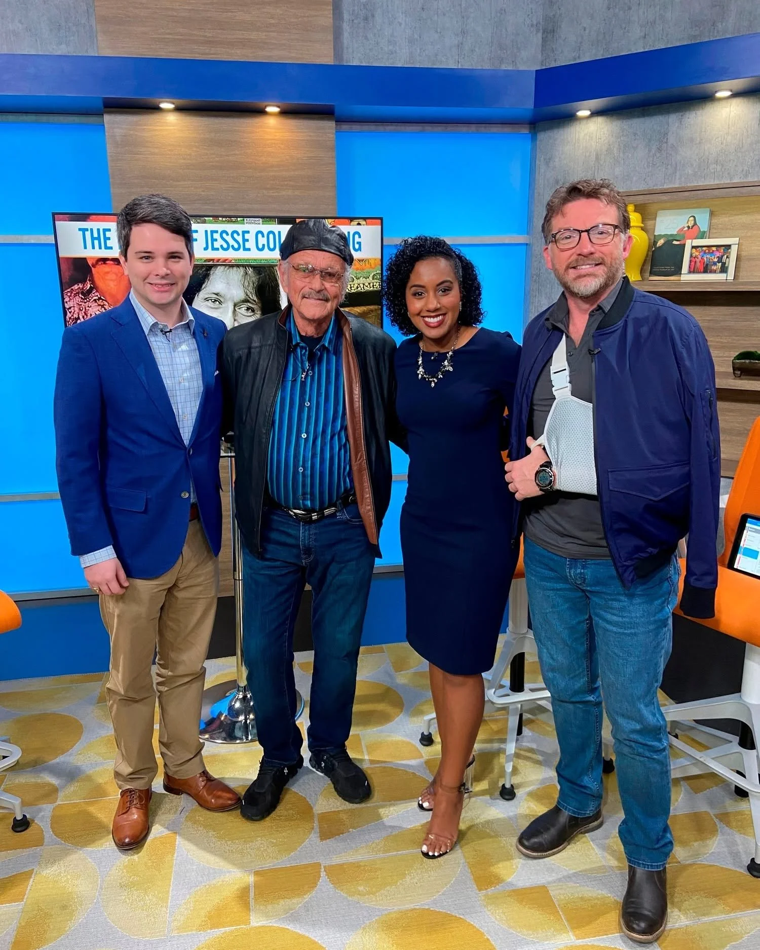 Four people standing in a studio, smiling for a photo, with a television behind them displaying a sign that says 'The Jesse Col...' and decorative shelves on the wall.