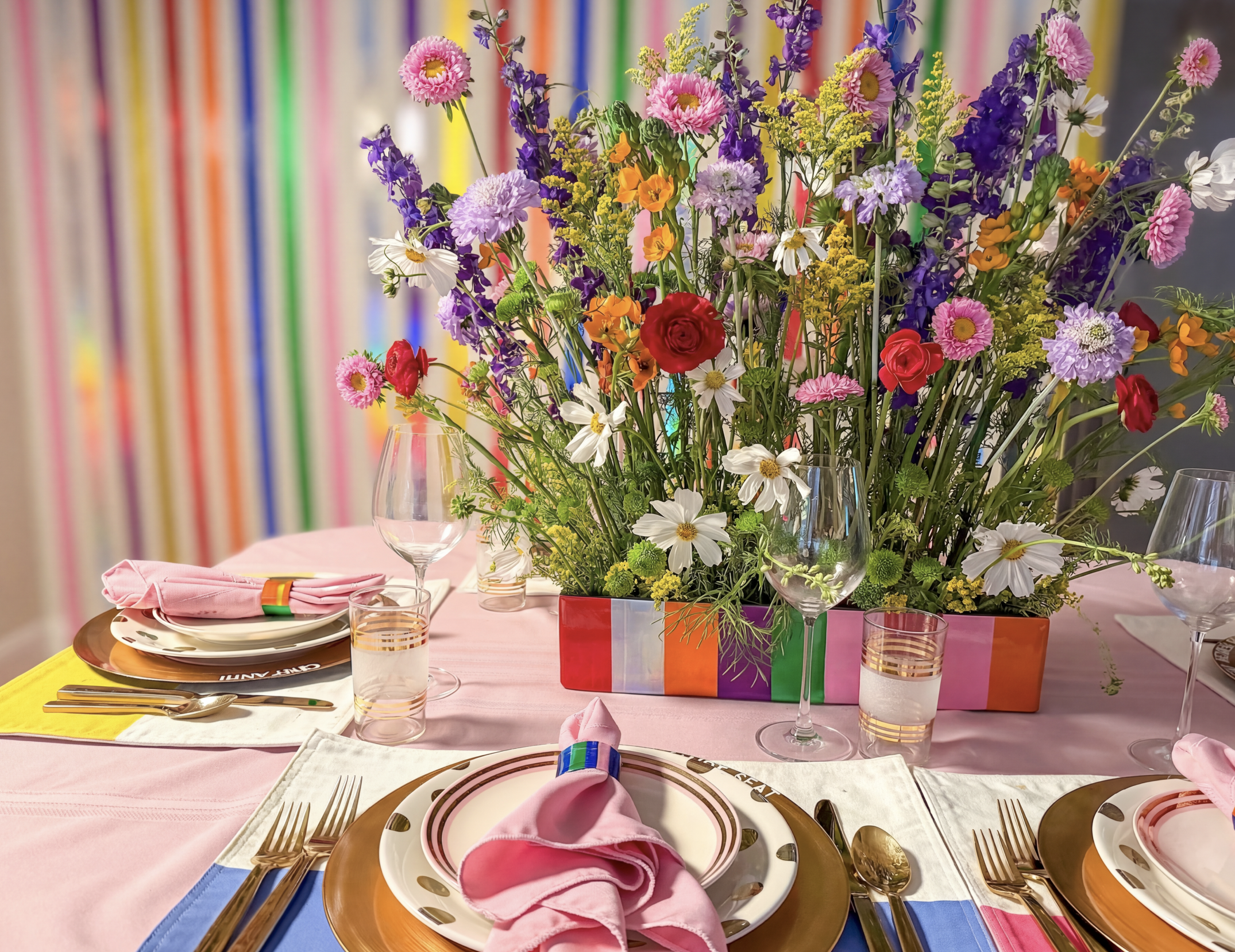 A colorful flower centerpiece on a pink tablecloth at a celebration or party table setting. The background features multicolored vertical stripes, adding a festive atmosphere.