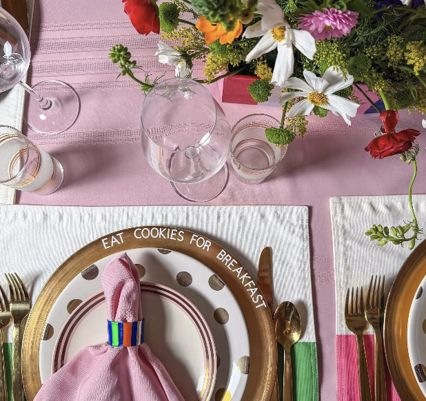 A colorful table setting with floral centerpiece, gold and white plates, pink cloth napkin, and green and pink utensils on a pink tablecloth.