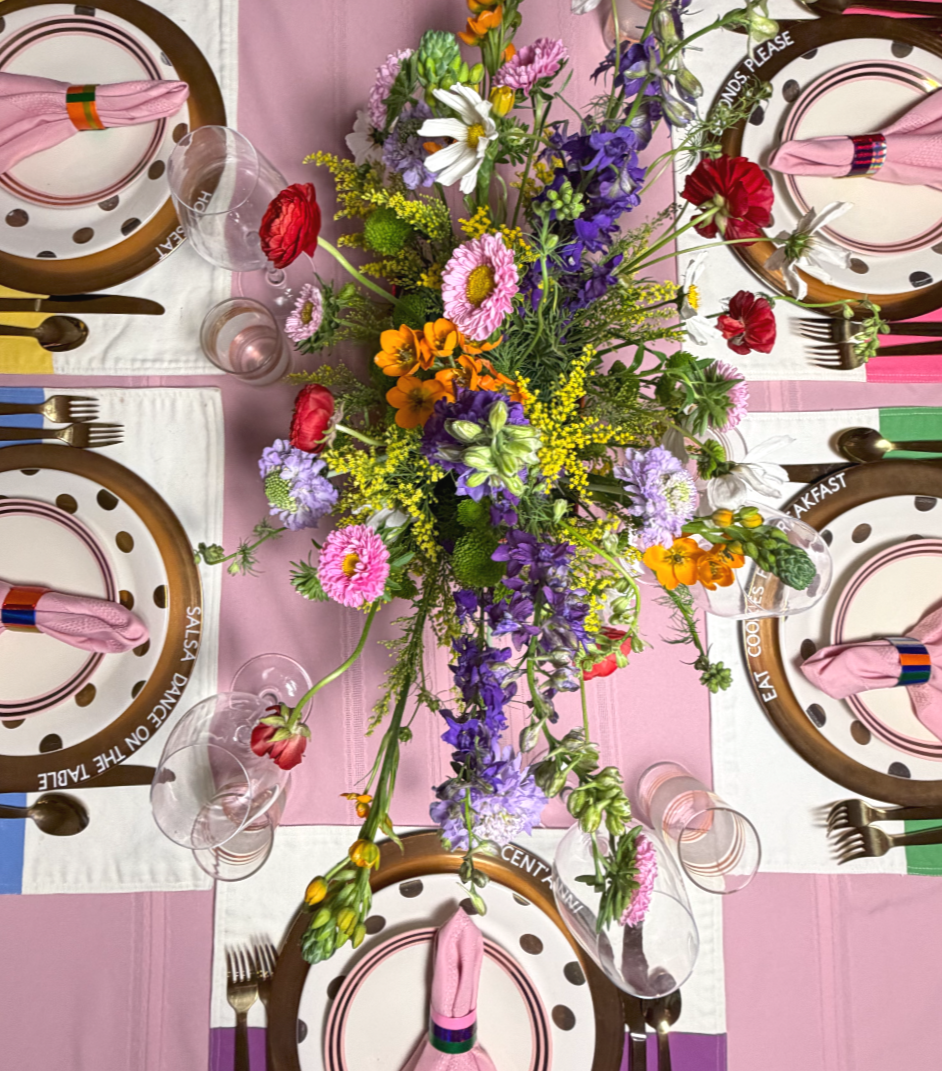 Table setting with floral centerpiece, pink tablecloth, white placemats with brown borders, pink napkins, and glassware for a celebration or gathering.