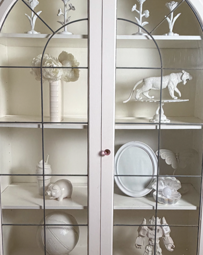 White display cabinet with glass doors containing decorative objects including a porcelain dog figurine, a ceramic flower bouquet in a vase, a toy robot, a globe, and various other white decorative items.