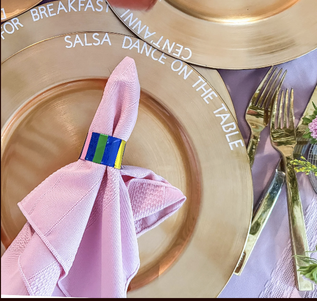 A pink cloth napkin folded and secured with a multicolored napkin ring, placed on a gold charger plate. The plate has white text around its edge that says 'Breakfast, Salsa Dance on the Table, Cyn-Fanny.' To the right, there are two gold forks and part of a purple tablecloth with a floral arrangement.