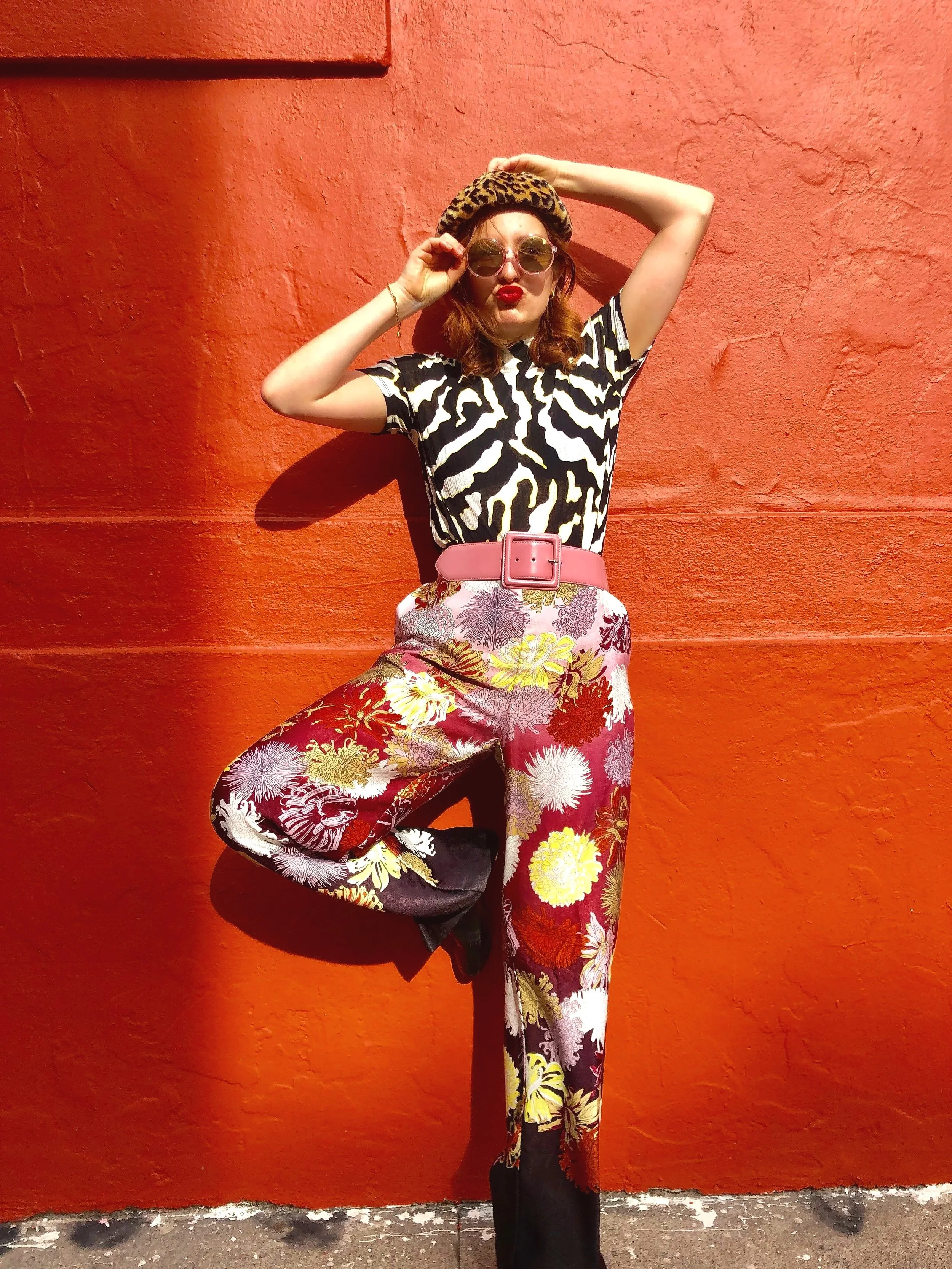A woman standing against a bright orange wall, wearing a zebra print shirt, floral pants, a pink belt, leopard print beret, sunglasses, and red lipstick, striking a pose with one hand on her head and the other adjusting her sunglasses.