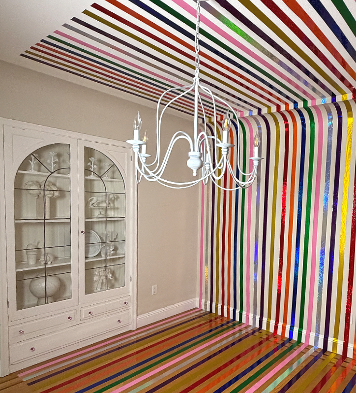 Room with colorful striped wallpaper on the walls and ceiling, a white chandelier hanging from the ceiling, and a white cabinet with glass doors displaying dishes.
