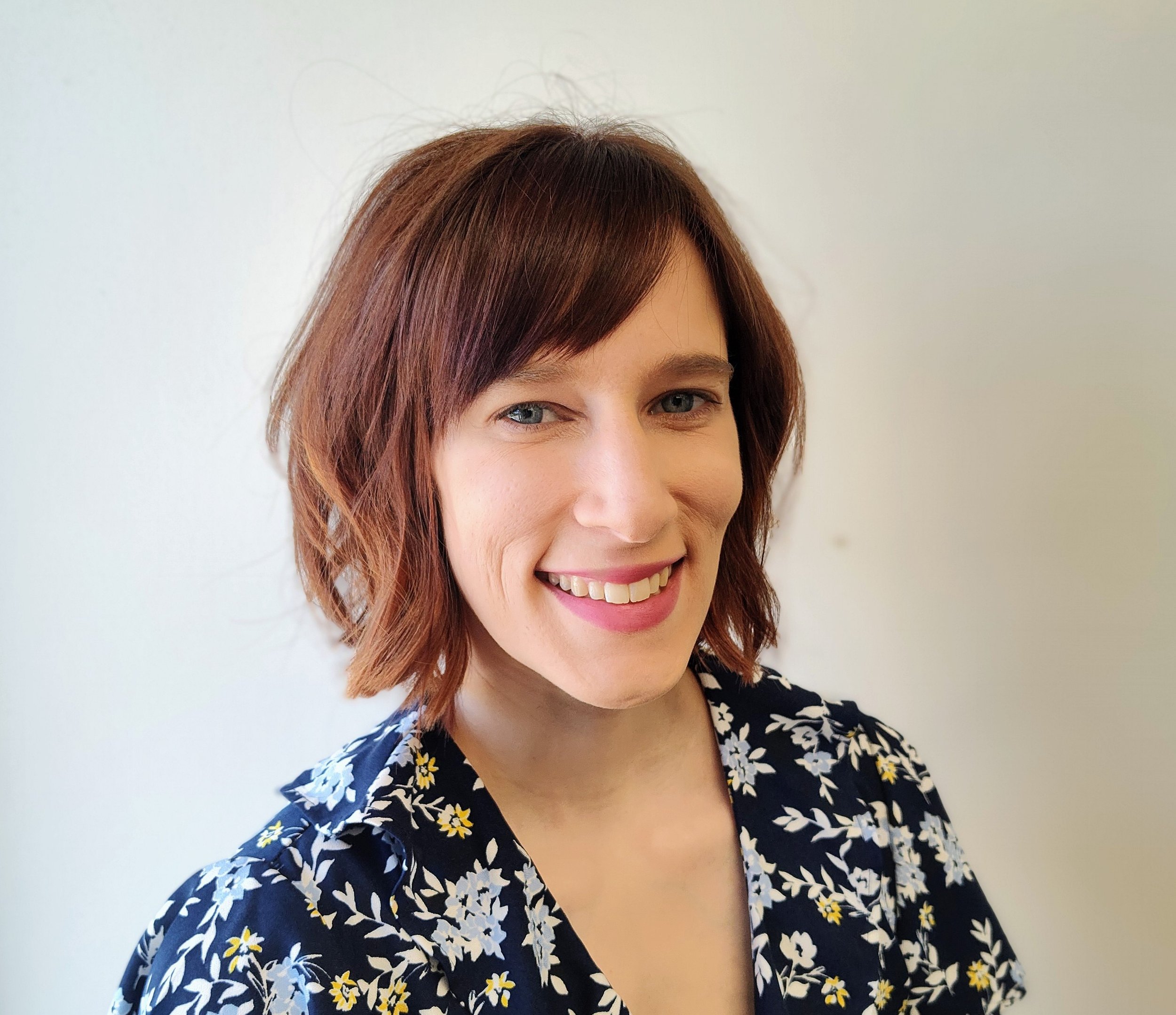 Close-up of a smiling woman with red hair, wearing a dark floral-patterned top, standing against a plain white wall.