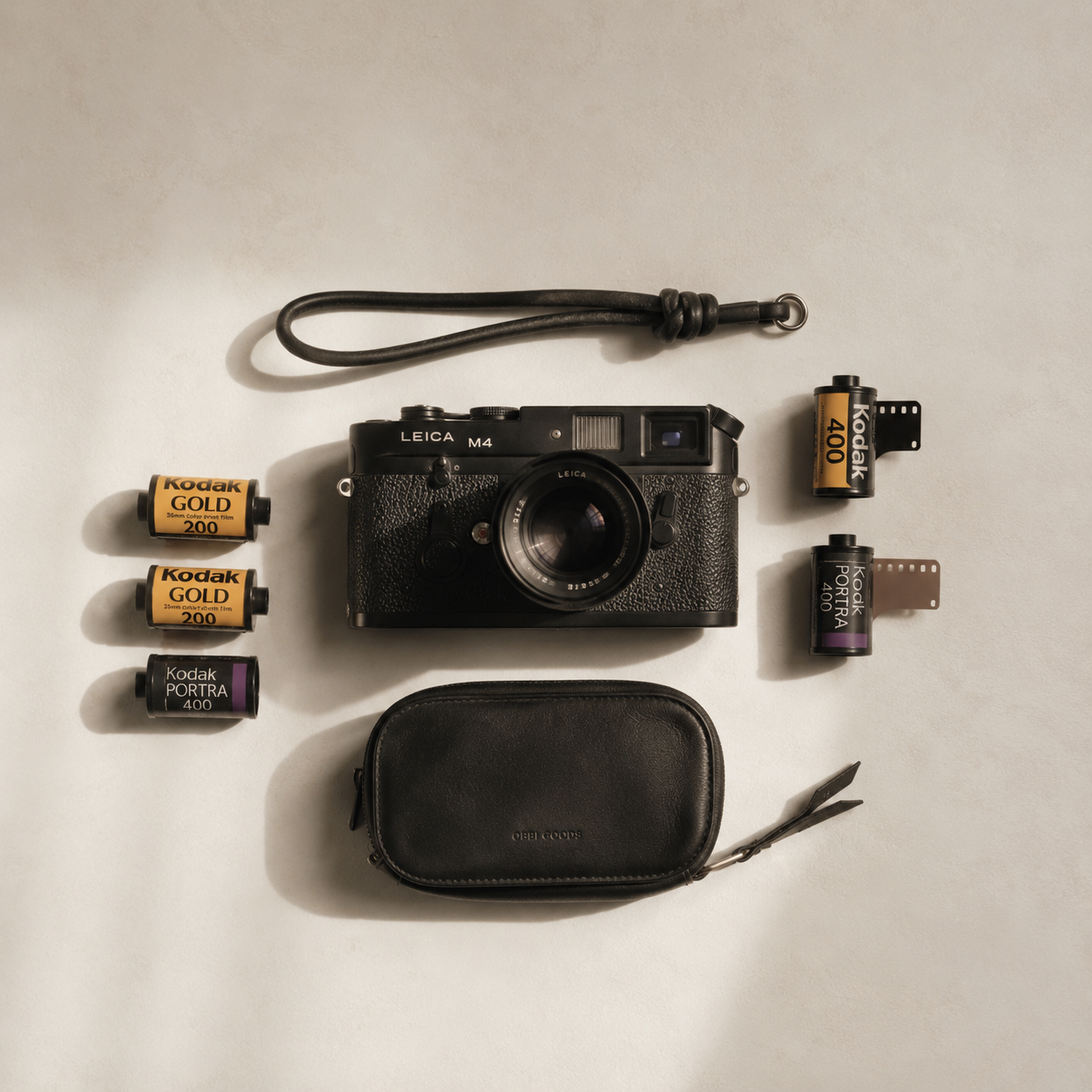 Eine Leica M4 Kamera, mehrere Kodak Filmrollen, ein schwarzes Kamerafutteral, ein Lichtzubehör und ein kleines Schneidewerkzeug auf einem weißen Hintergrund.