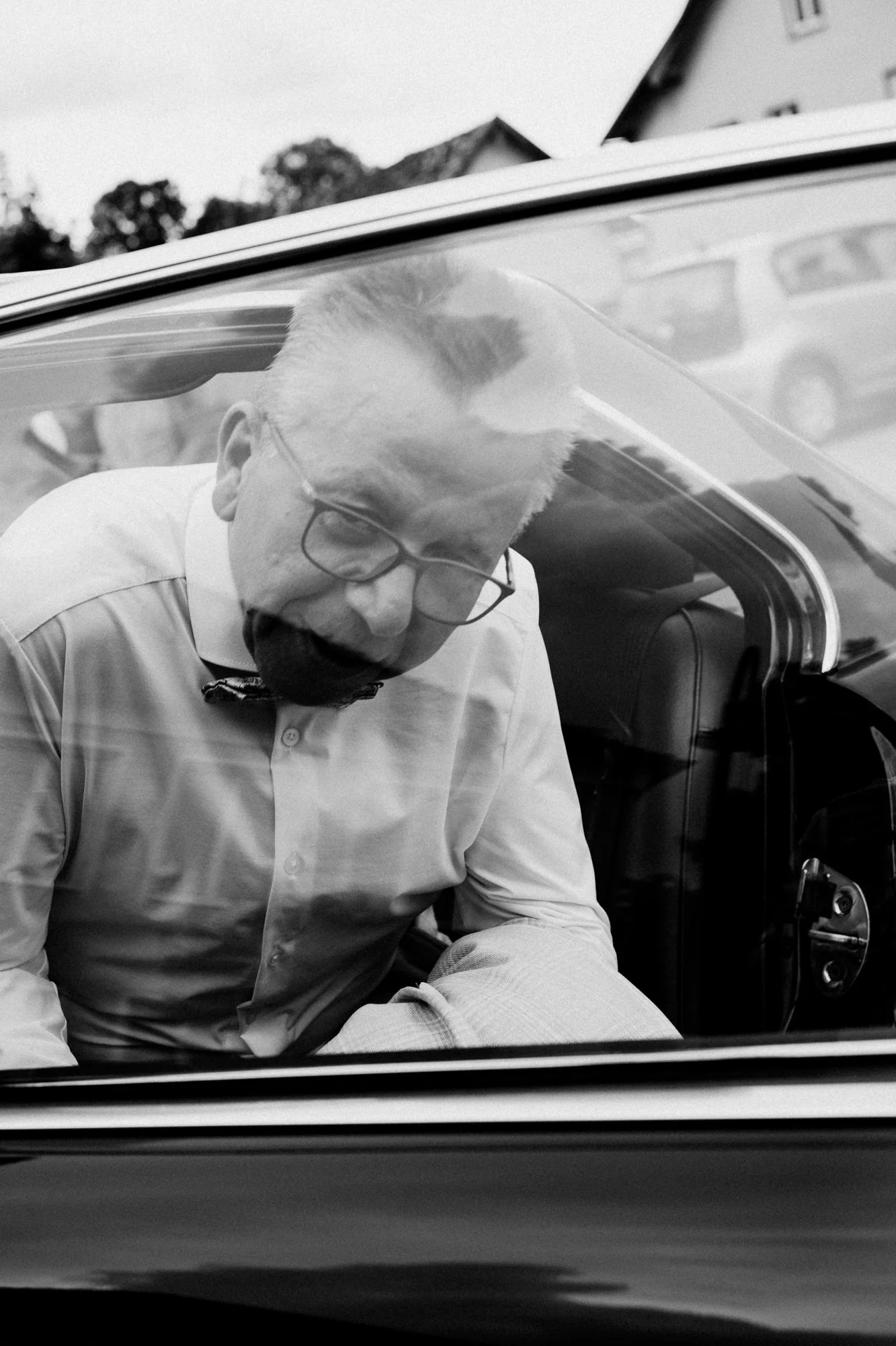 Schwarz-weiß-Foto von einem Mann mit Brille, der in einem Auto sitzt und durch die offene Tür einen Blick nach innen wirft.