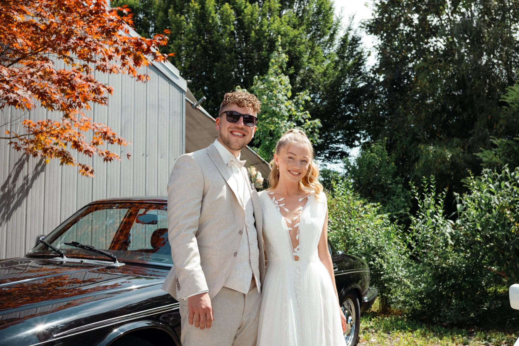 Ein glückliches Paar in formeller Kleidung, vermutlich auf einer Hochzeit, steht neben einem schwarzen Oldtimer-Auto im Grünen.