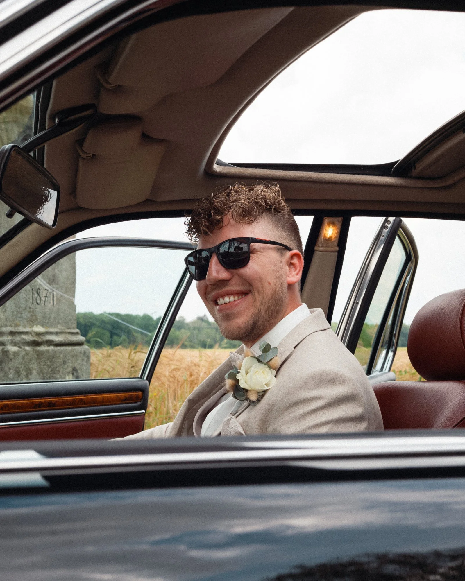 Junger Mann in einem Hochzeitsanzug sitzt in einem Auto, trägt Brille und hat einen Blumenkranz am Revers. Im Hintergrund ist eine ländliche Landschaft zu sehen.