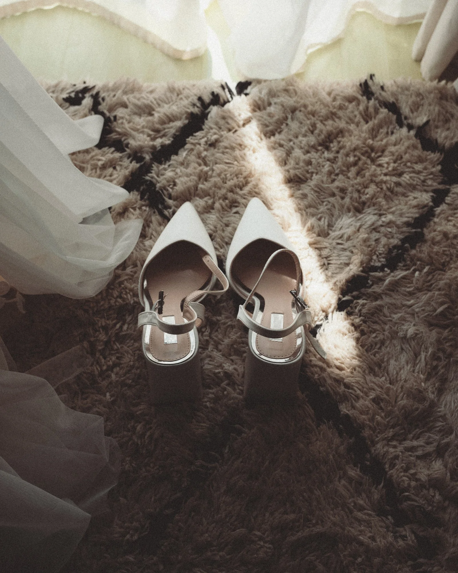 Ein Paar weiße High Heels auf einem flauschigen Teppich, daneben eine weiße Einkaufstüte, im Hintergrund eine Tür und eine Grünwand.