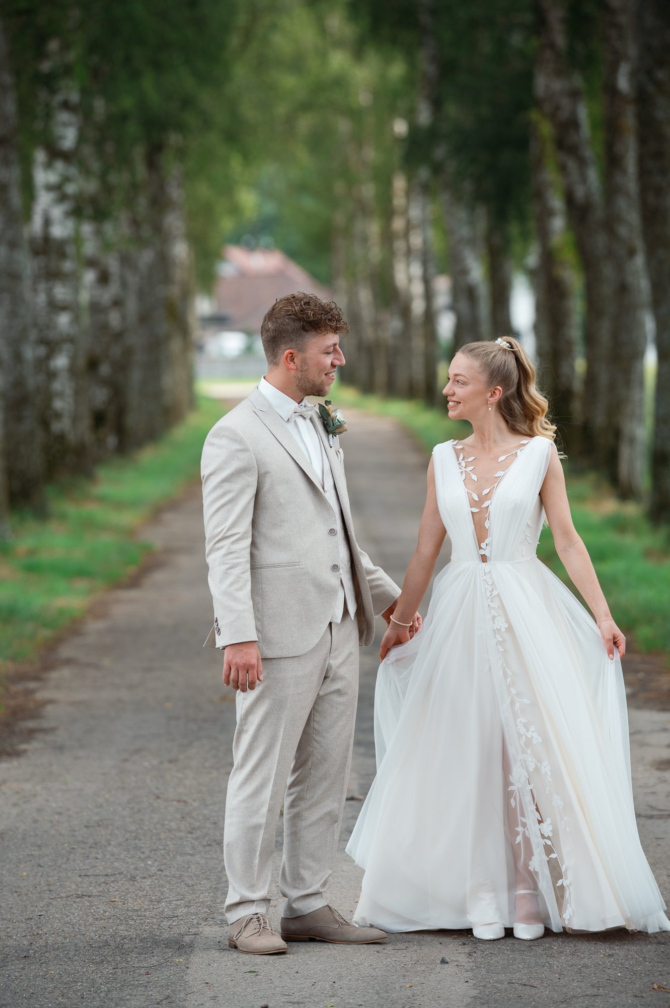 Ein verliebtes Paar bei einer Hochzeit, die auf einer Allee mit Bäumen steht, hält Händchen und schaut sich liebevoll an.