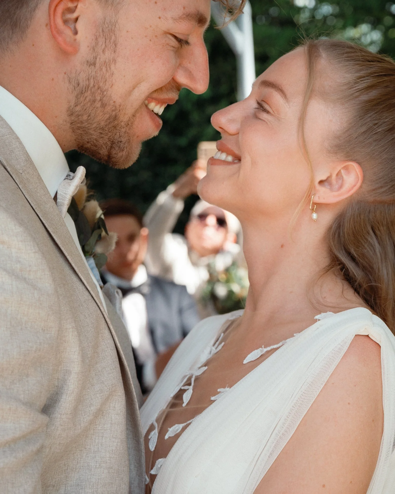 Ein Heiratsantrag zwischen einem Mann und einer Frau, beide lächelnd und liebevoll Blickkontakt, im Freien bei Sonnenuntergang, mit Freunden im Hintergrund.