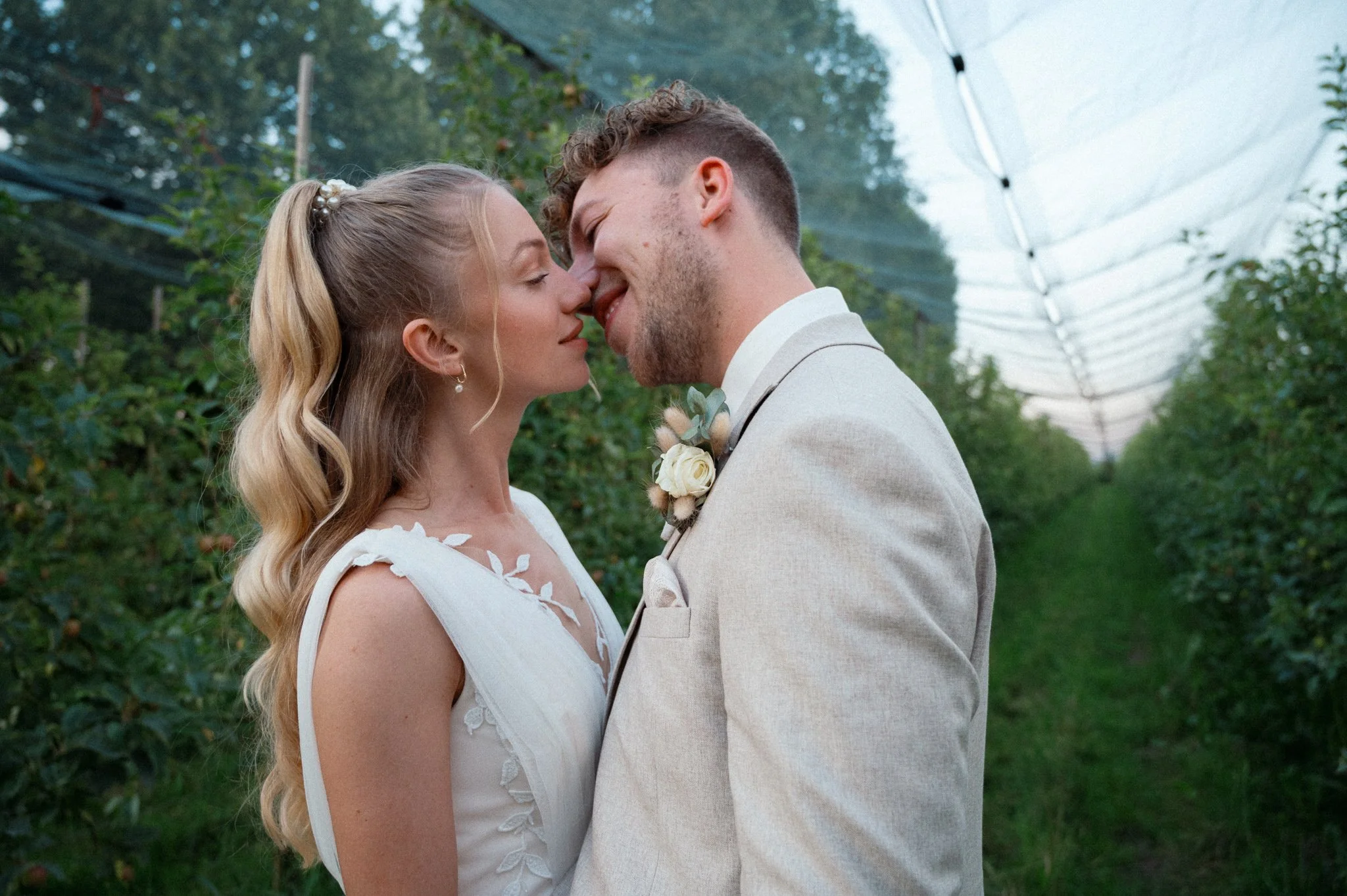 Ein verliebtes Paar bei einer Hochzeit steht sich mit der Stirn aneinander, in einer grünen, bewachsenen Umgebung unter einem weißen Tunnel.