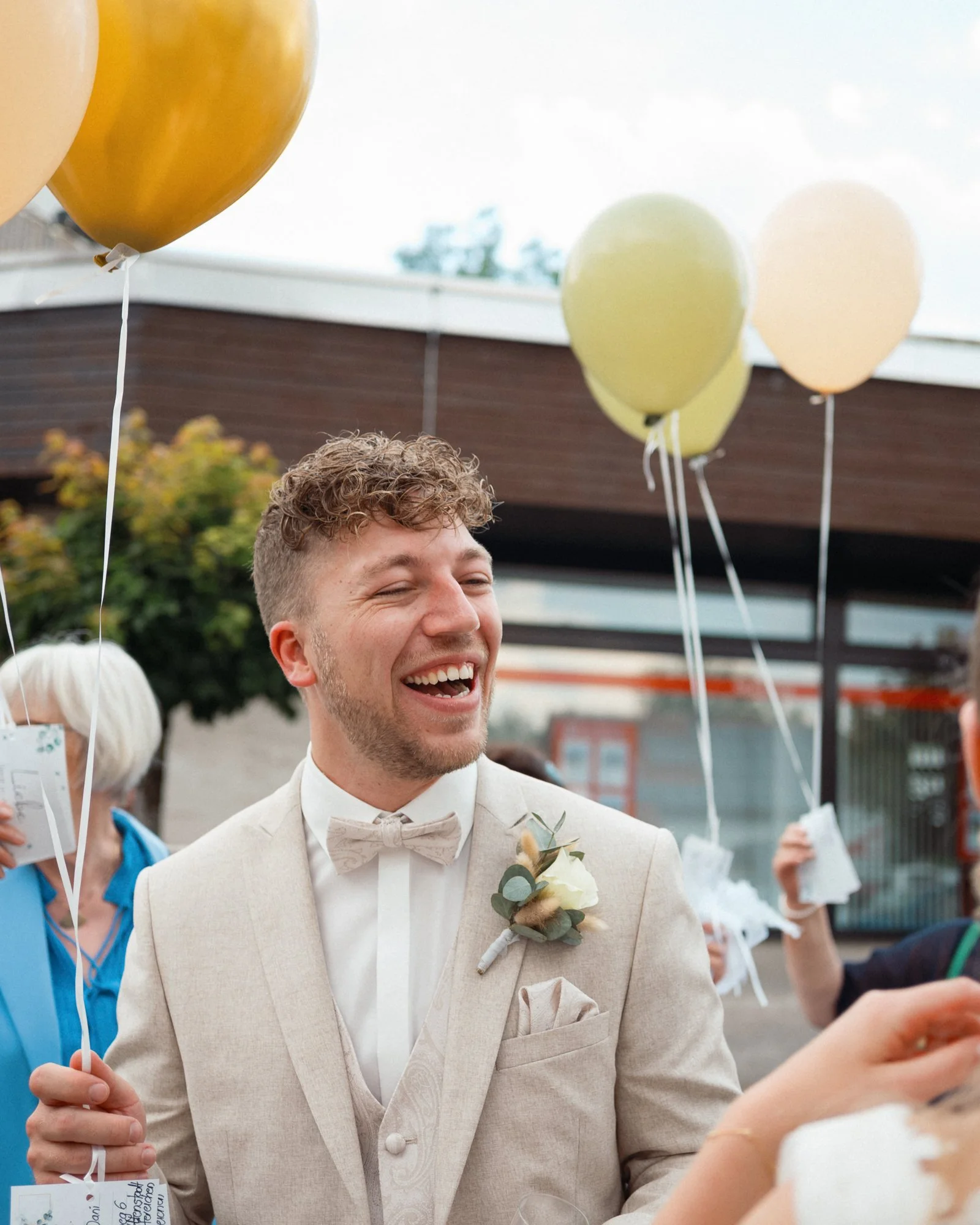 Ein lachender Mann in einem cremefarbenen Anzug mit Fliege bei einer Hochzeit, umgeben von heliumgefüllten Ballons, während er ein Handy hält.